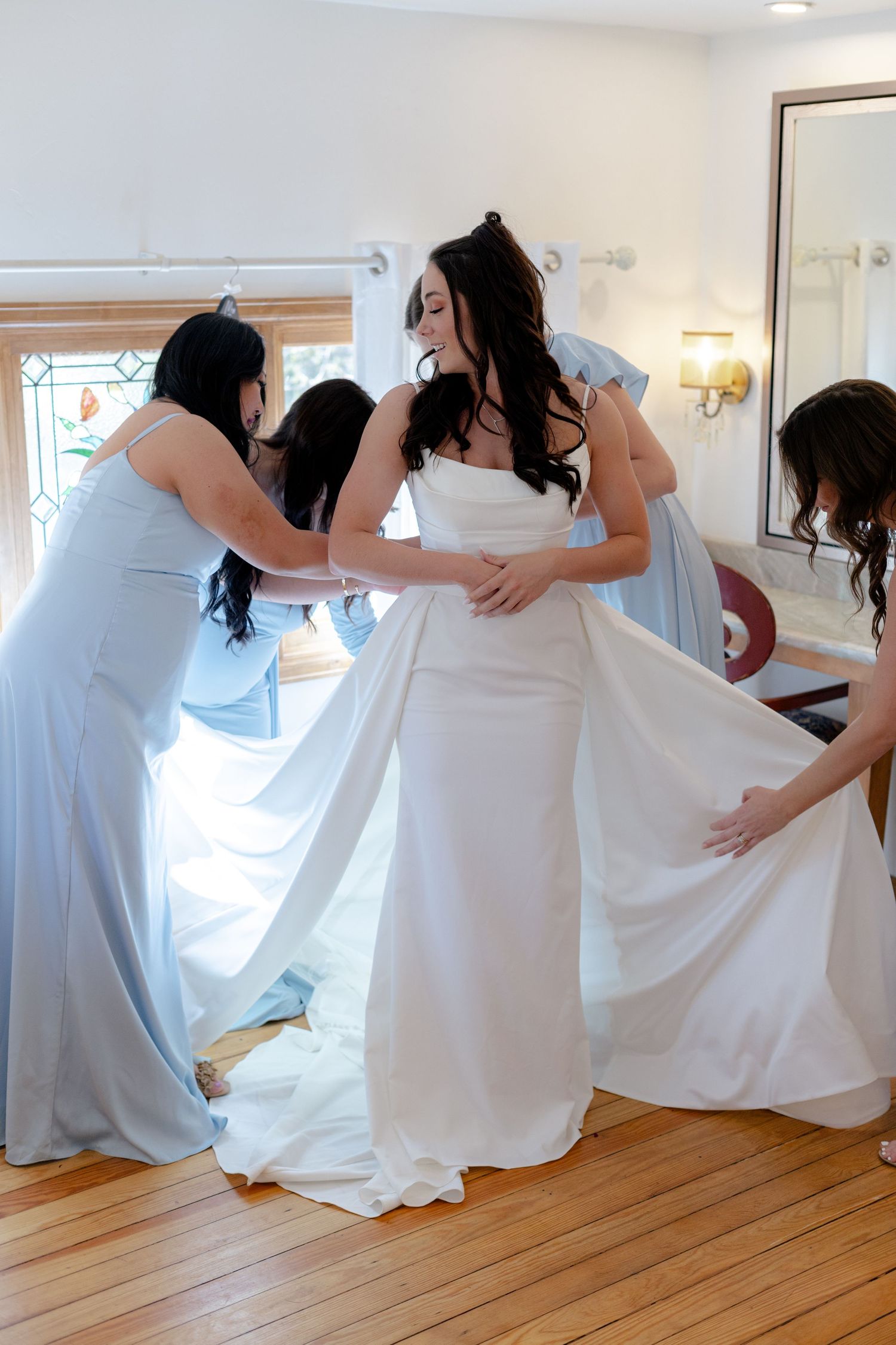 Bridesmaids in light blue dresses helping bride into elegant white wedding gown in bright dressing room.