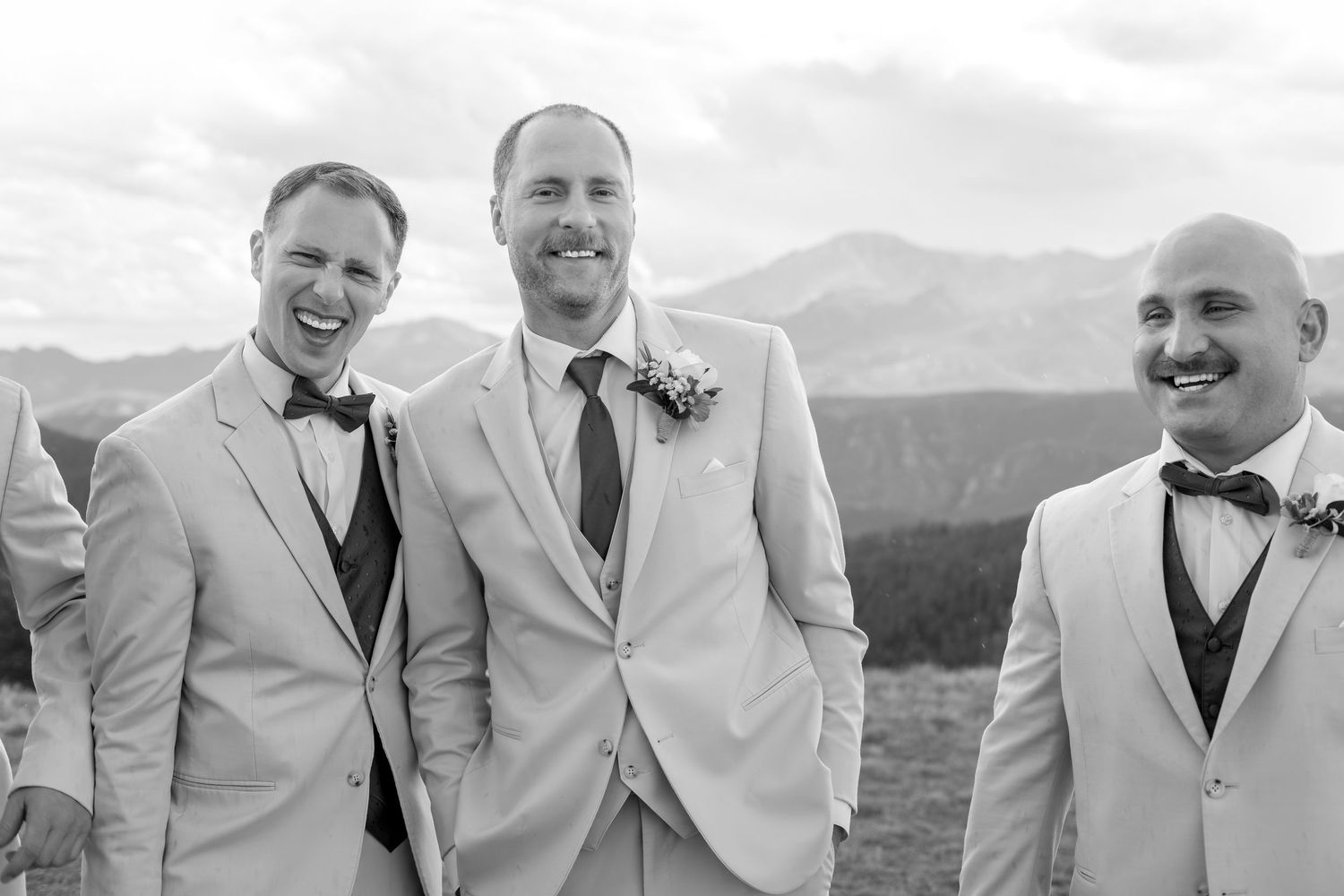 Three groomsmen in light suits and bow ties laughing outdoors with mountain scenery backdrop at a wedding.