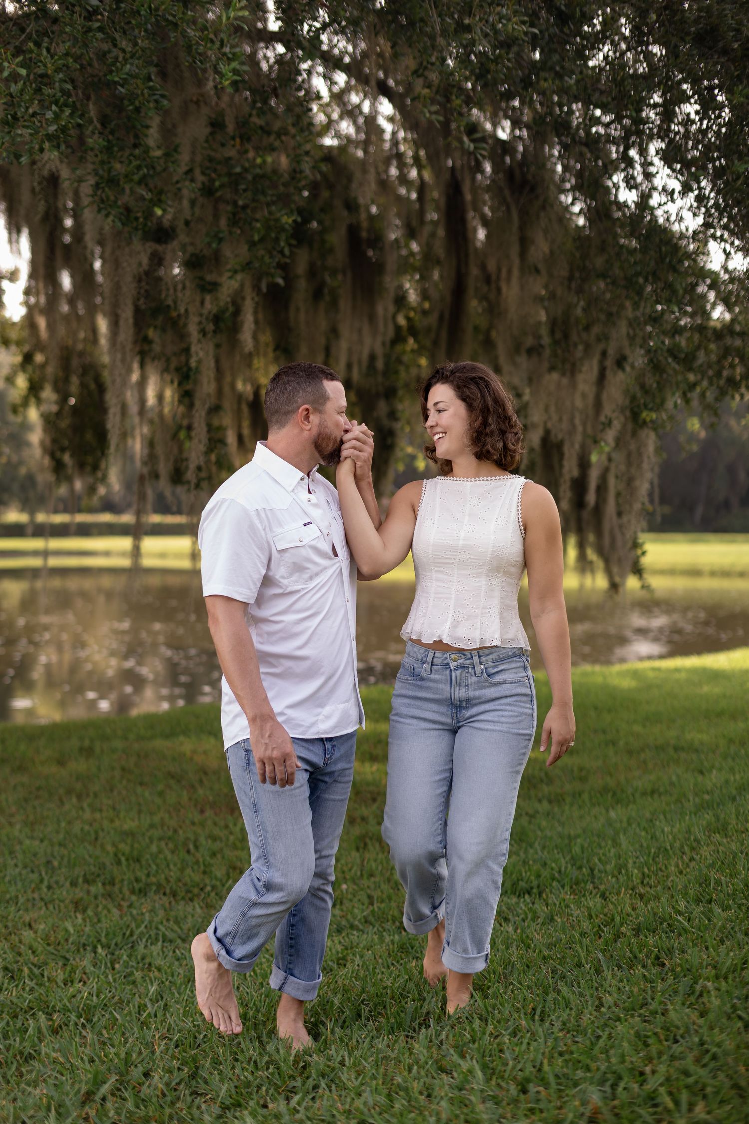 Couple walks barefoot on green grass, man kissing woman's hand near moss-draped oak trees by a pond.