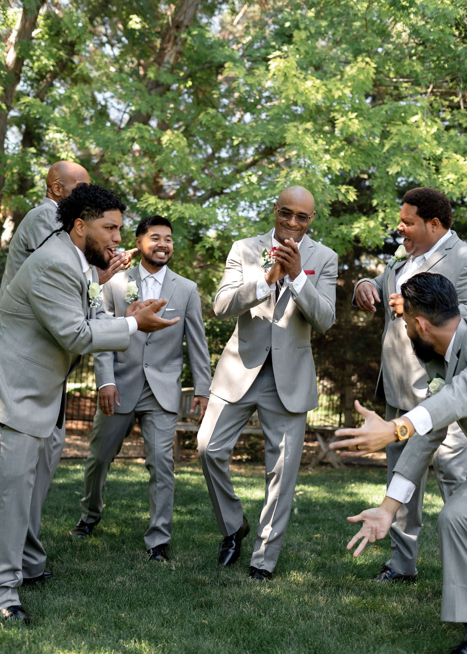 Groomsmen in gray suits laughing and celebrating outdoors at a wedding reception on a sunny day.