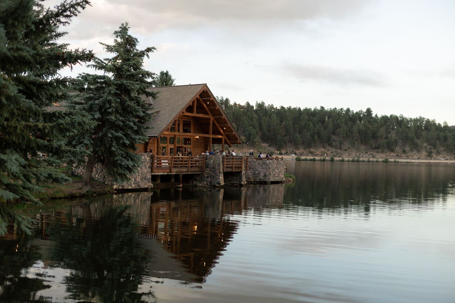 Rustic wooden cabin on stilts reflects in calm lake surrounded by dense pine forest under cloudy sky.