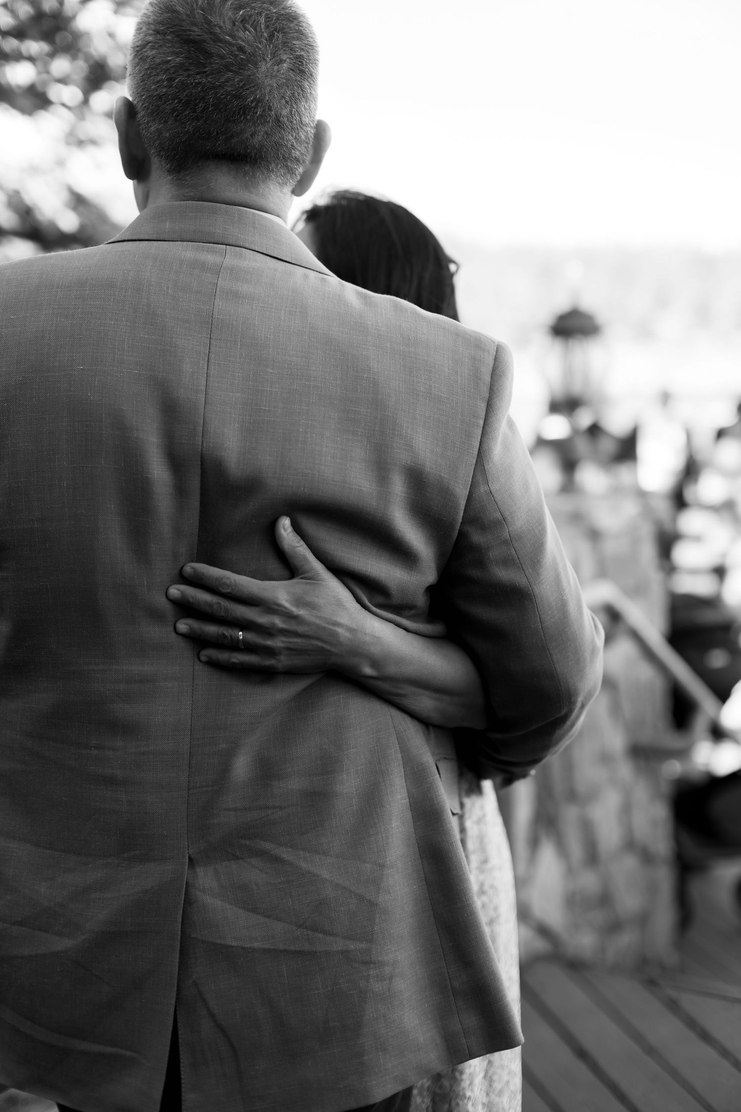 Black and white photo of a couple embracing at a wedding, woman's hand with ring resting on man's suited back.