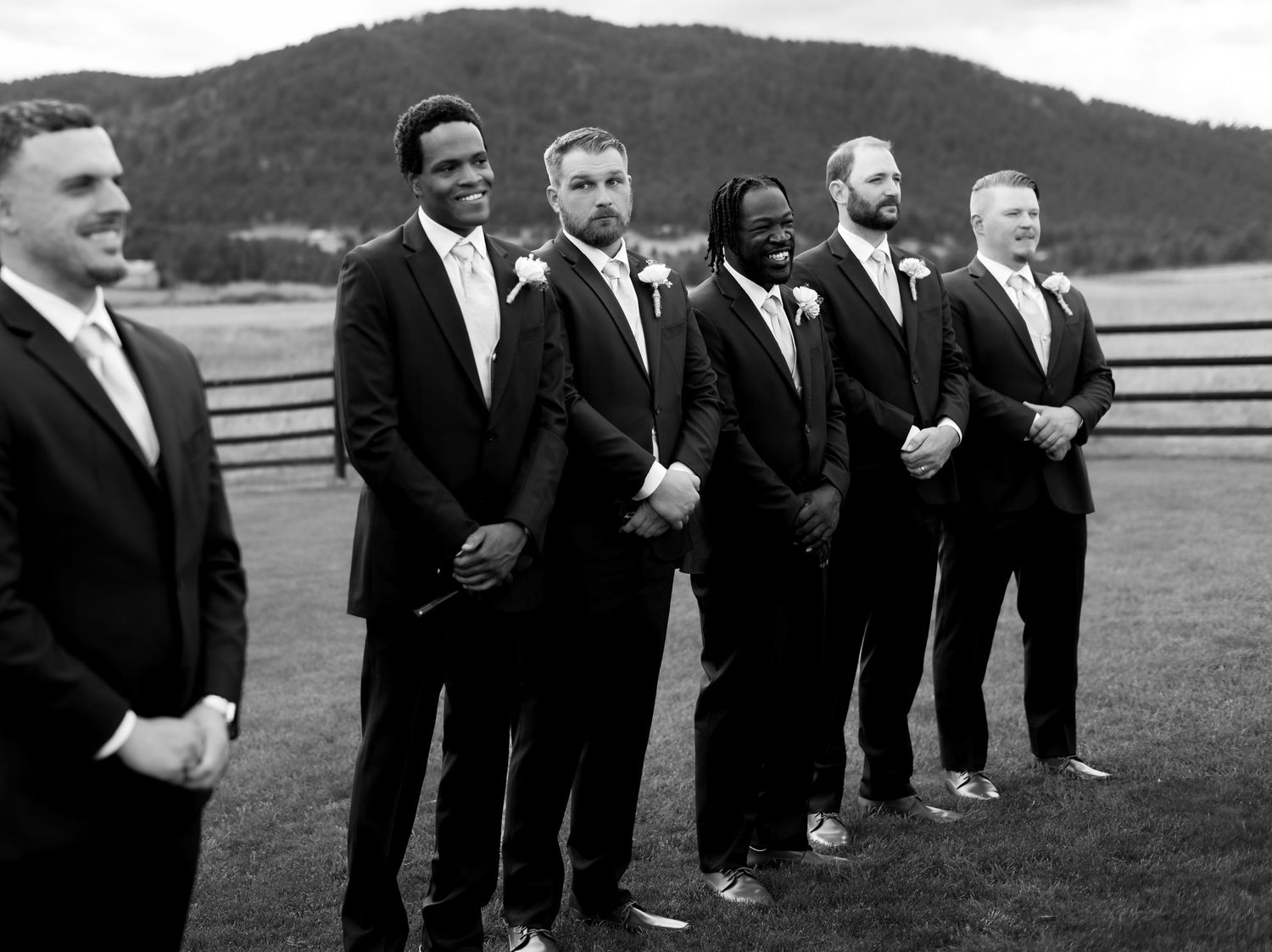 Black and white photo of six groomsmen in dark suits posing outdoors at a mountain wedding venue.