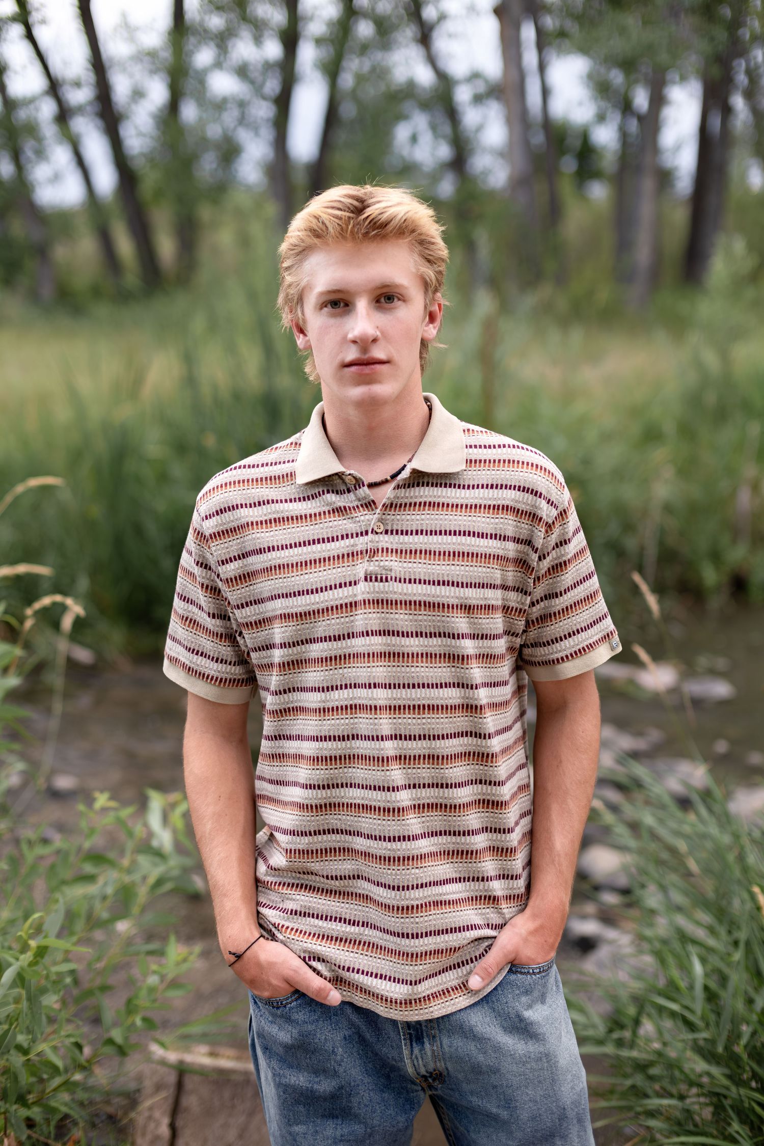 Young blond man in striped polo shirt and jeans standing outdoors with green trees and grass in background.
