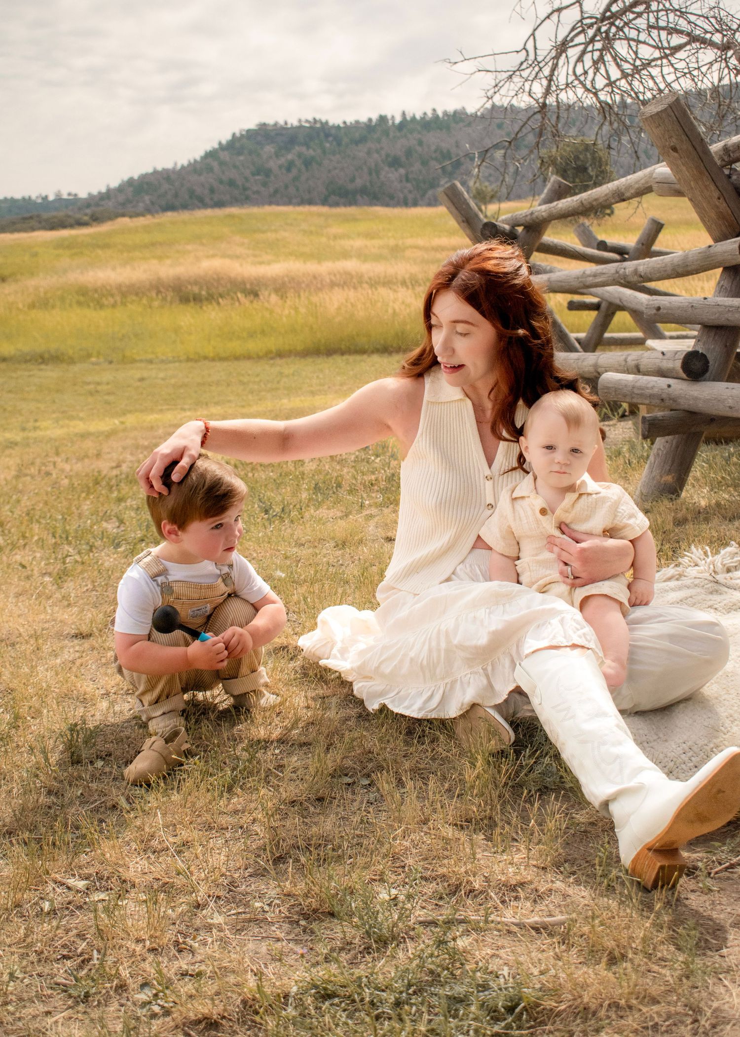 A red-haired woman in a white dress sits in a grassy field with two young children near a rustic wooden fence.