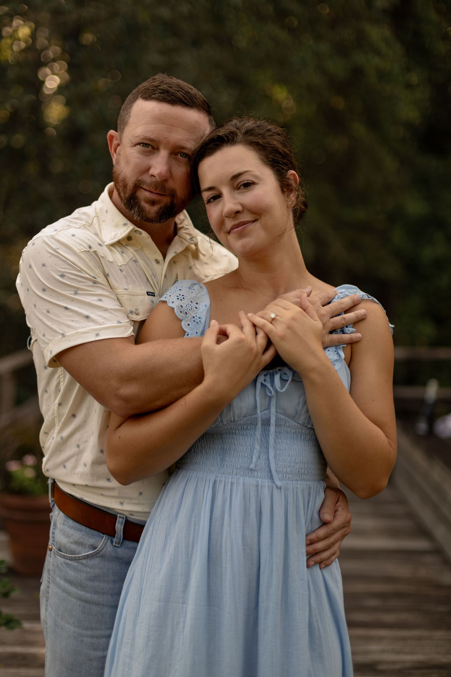 A couple embraces on a wooden bridge outdoors, woman in blue dress, man in white shirt and jeans, surrounded by greenery.