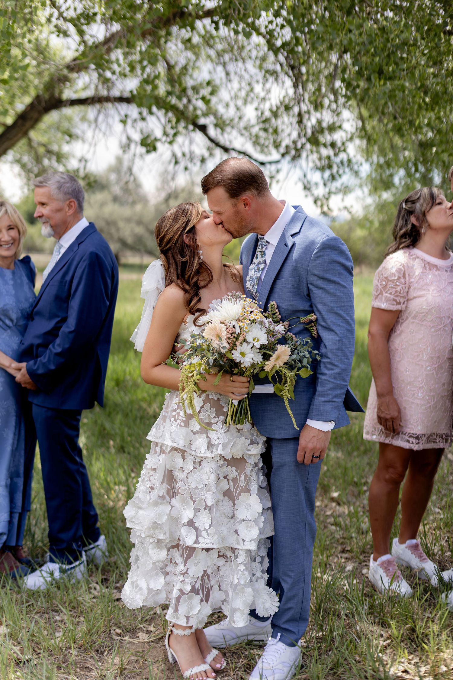 Bride and groom share a first kiss holding a wildflower bouquet with family smiling behind them.