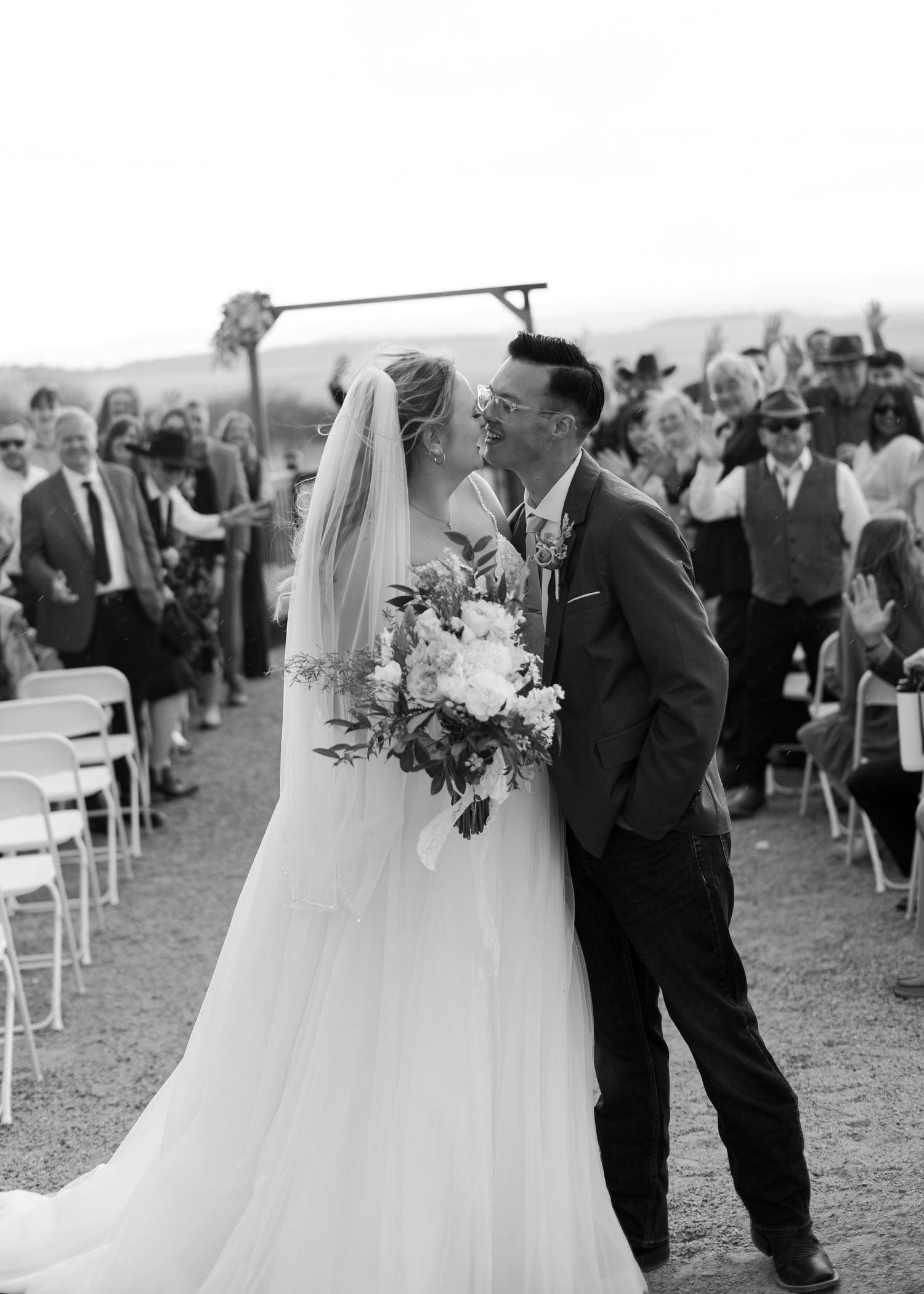 Black and white photo of a newlywed couple sharing a kiss at the altar while wedding guests cheer behind them.