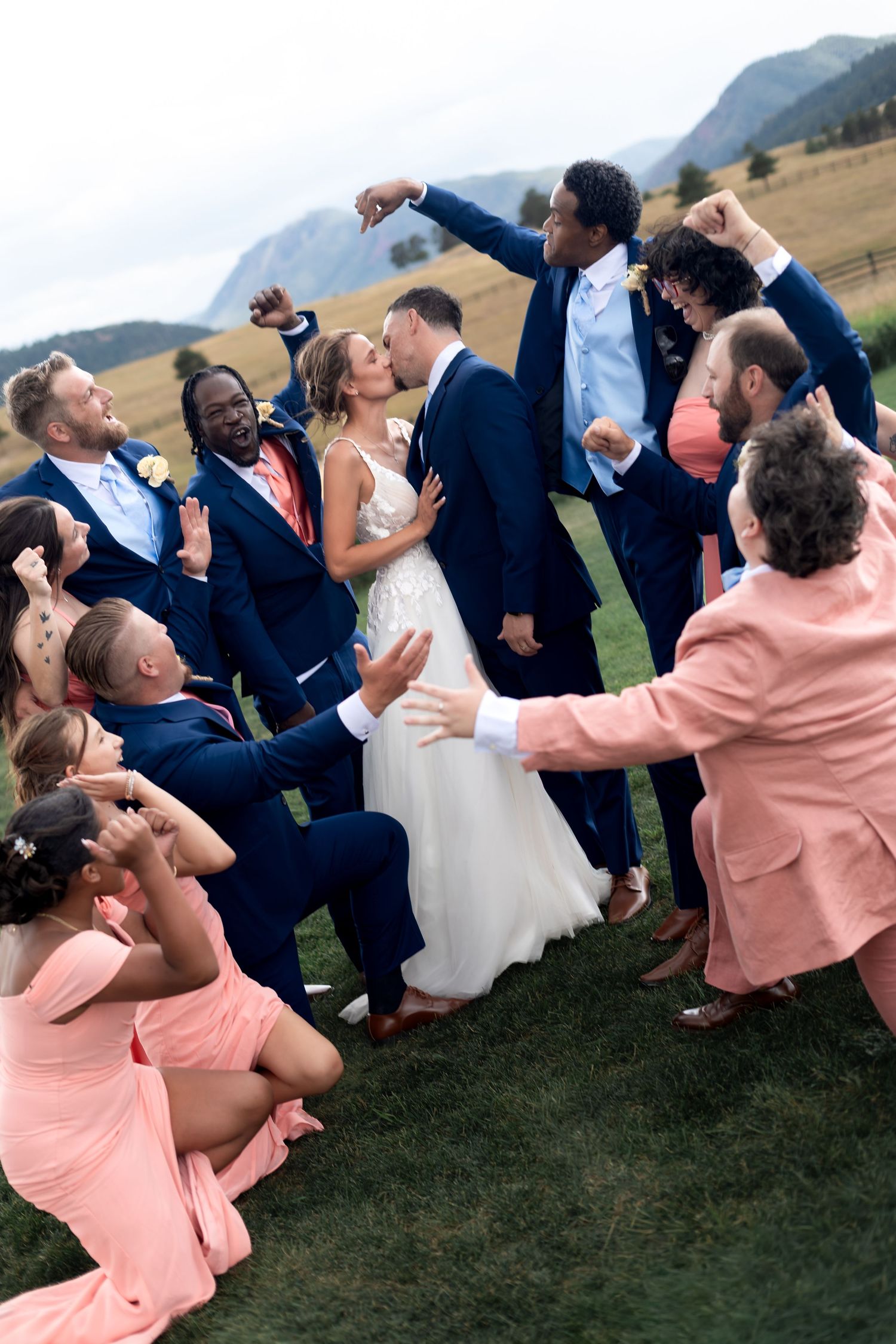 A joyful wedding party celebrates as the bride and groom share a kiss outdoors with mountains in the background.