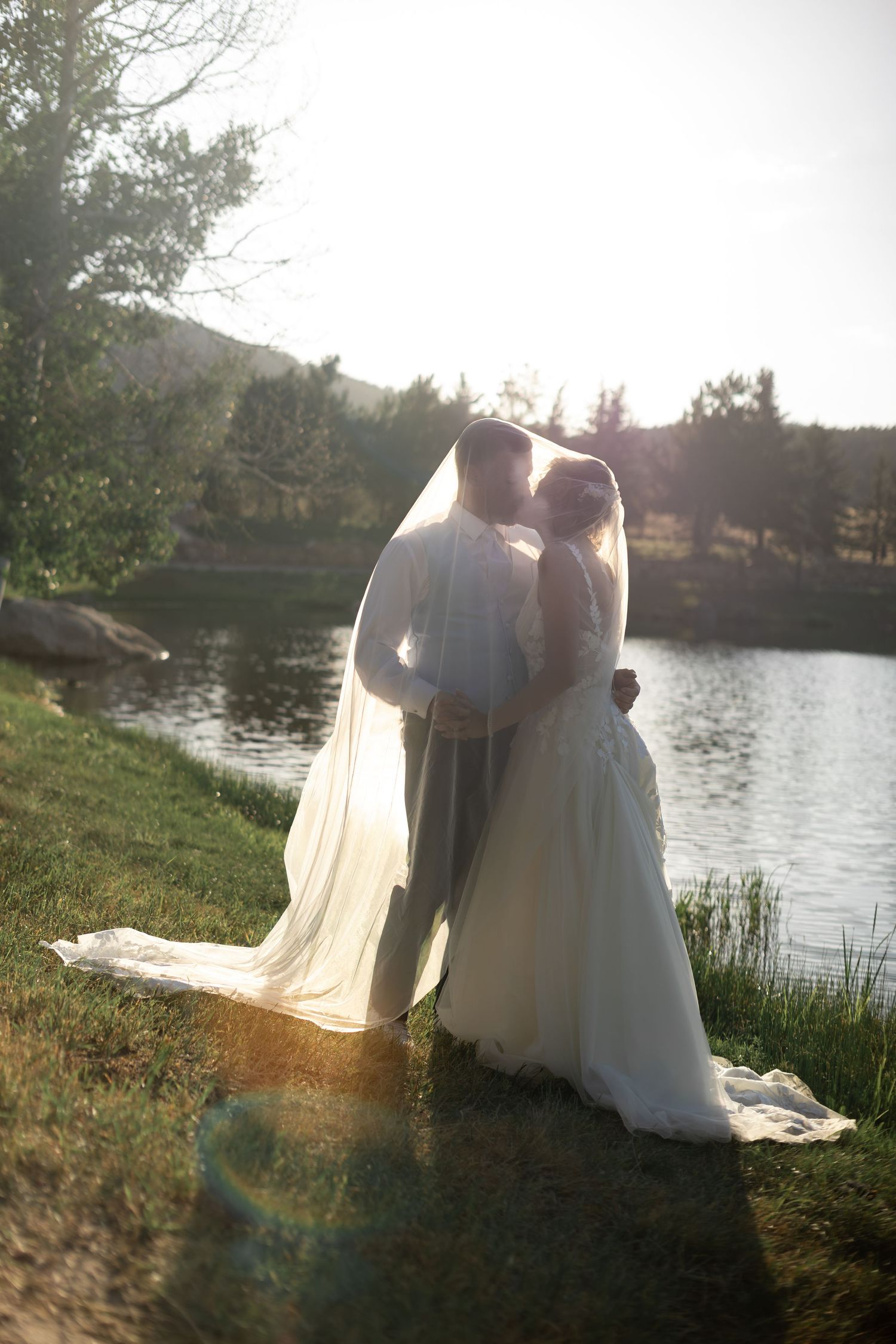 Bride and groom kissing by a sunlit lake, bride in white gown with flowing veil, surrounded by lush greenery.