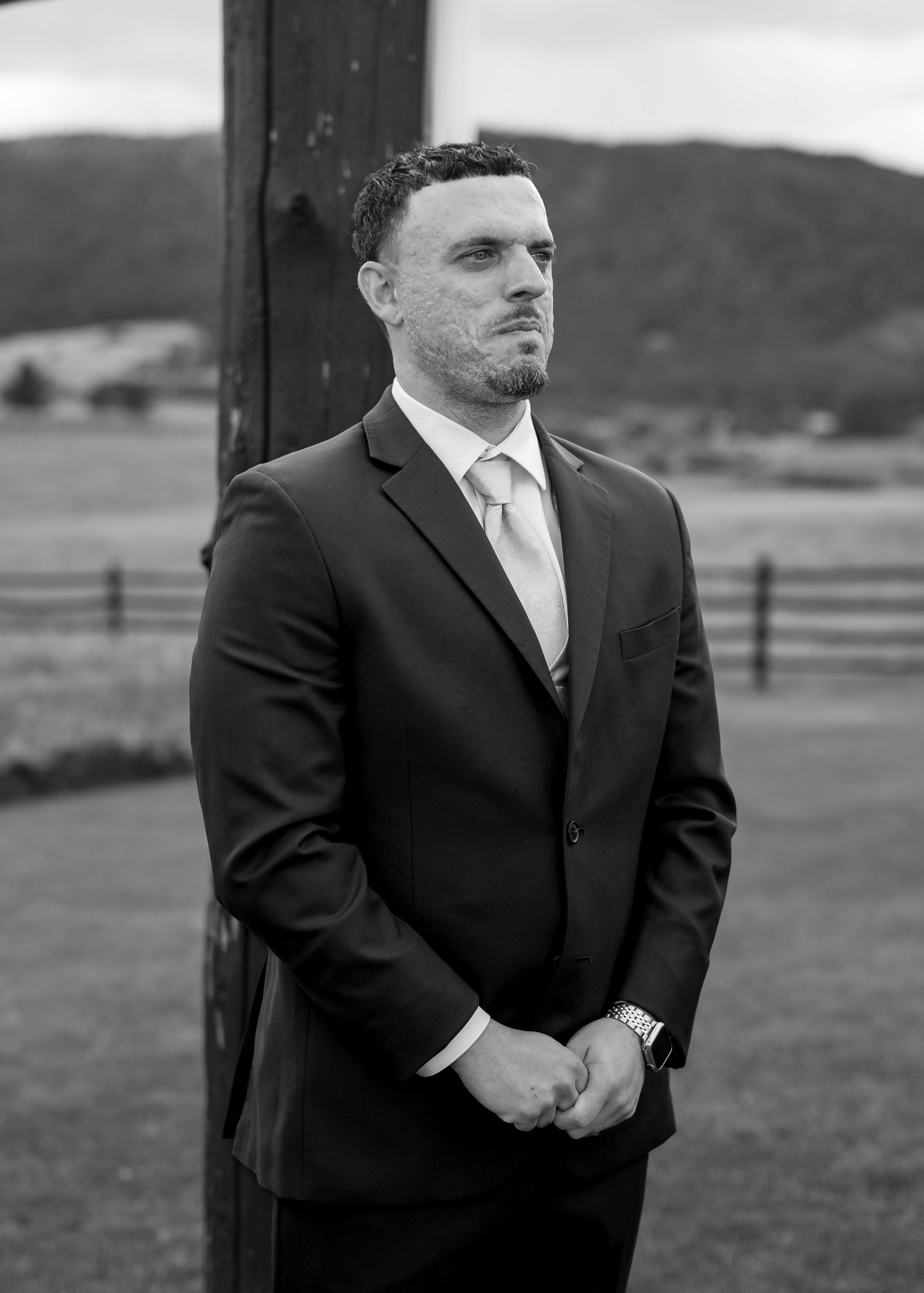 Groom in dark suit and light tie standing outdoors at a countryside wedding venue in black and white.