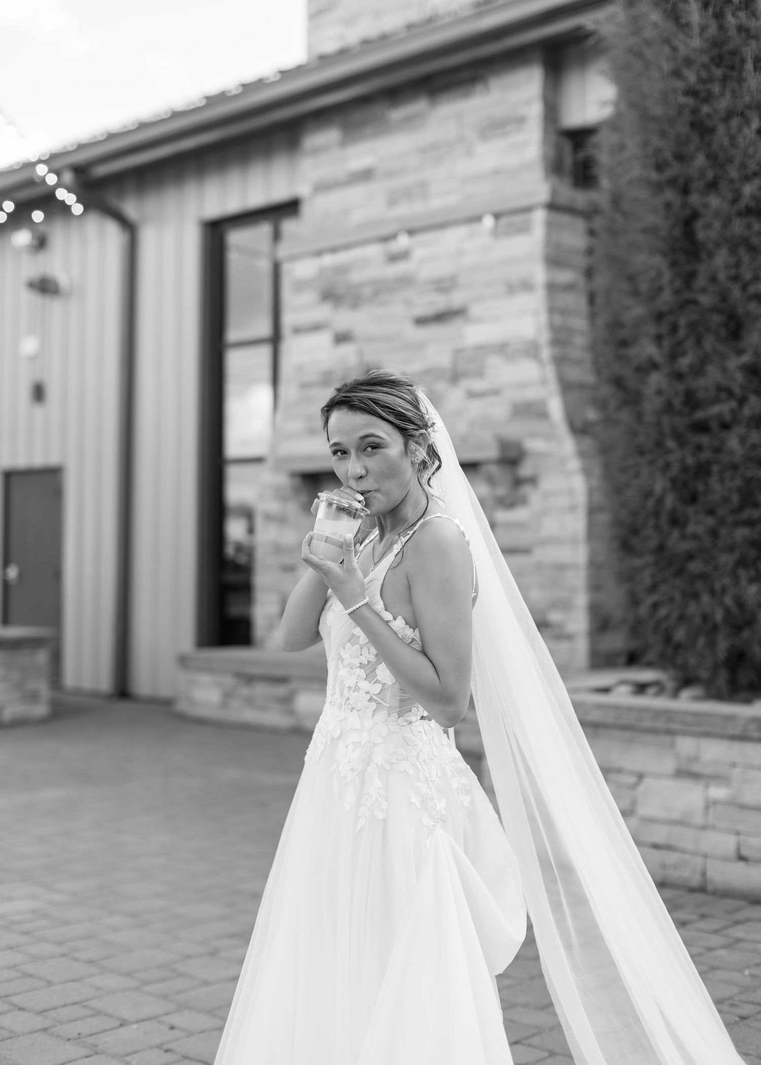 Bride in white lace gown and veil blows a kiss outside stone wedding venue in black and white.