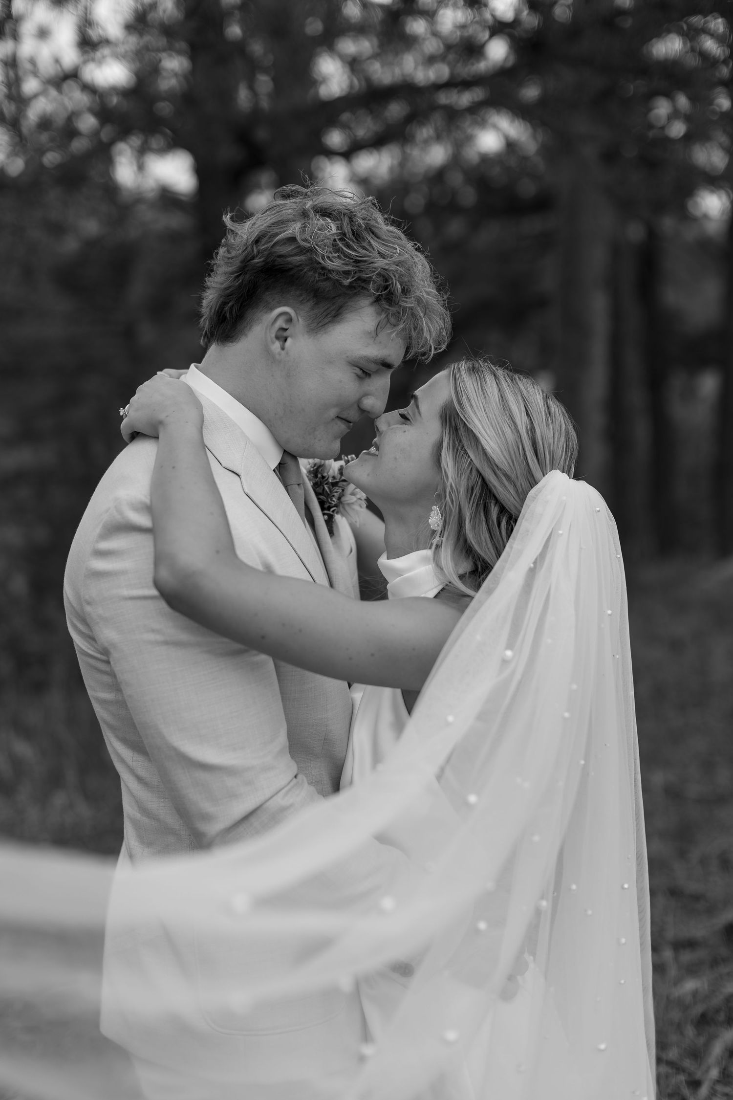 Black and white photo of bride and groom nearly kissing, veil flowing in the breeze among trees.