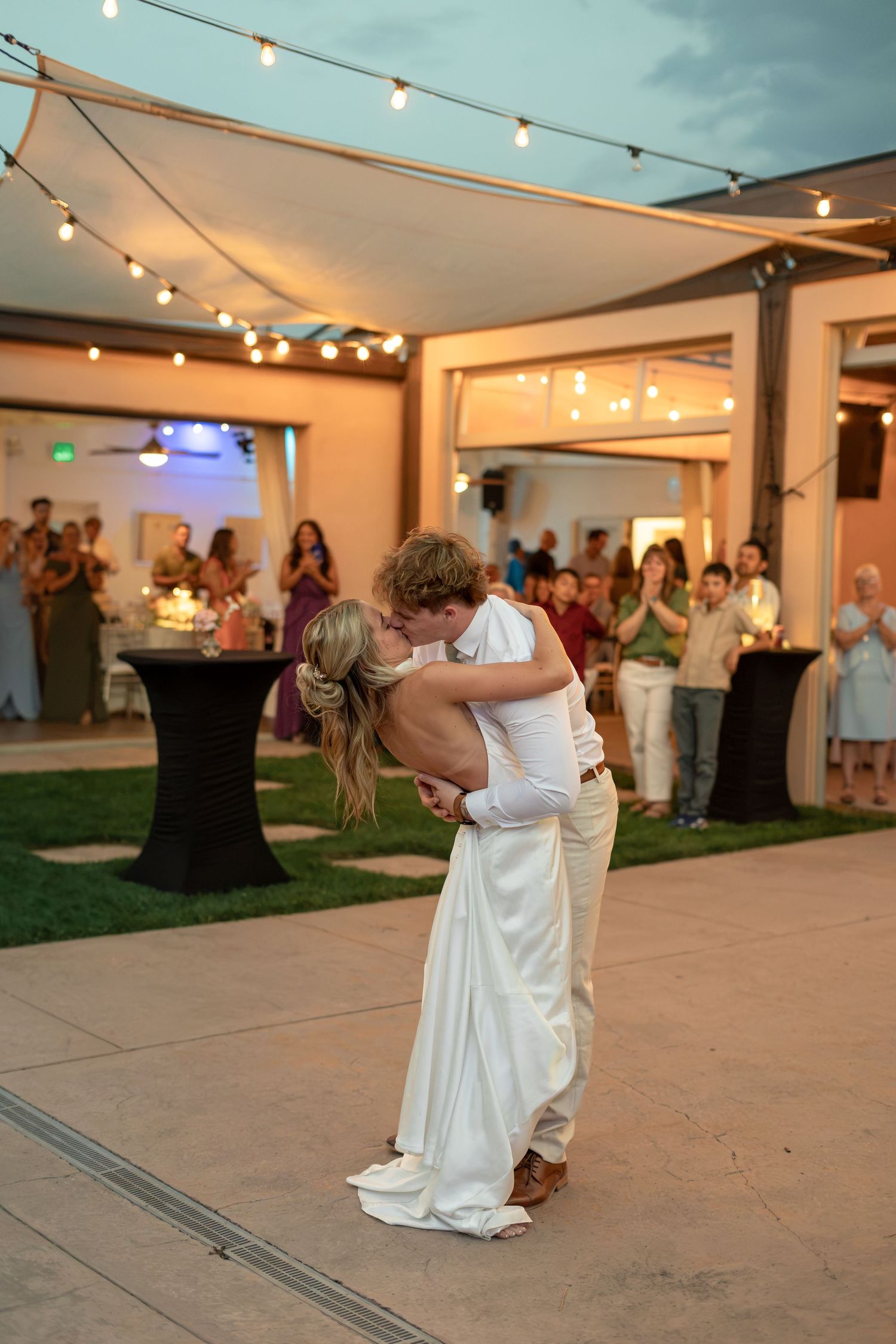 Bride and groom share romantic dip kiss on outdoor dance floor surrounded by string lights and wedding guests.