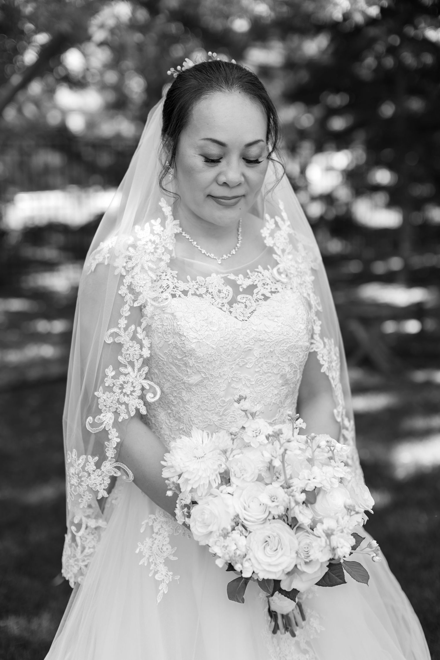 Bride in lace wedding dress and veil holds floral bouquet, looking down in black and white portrait.