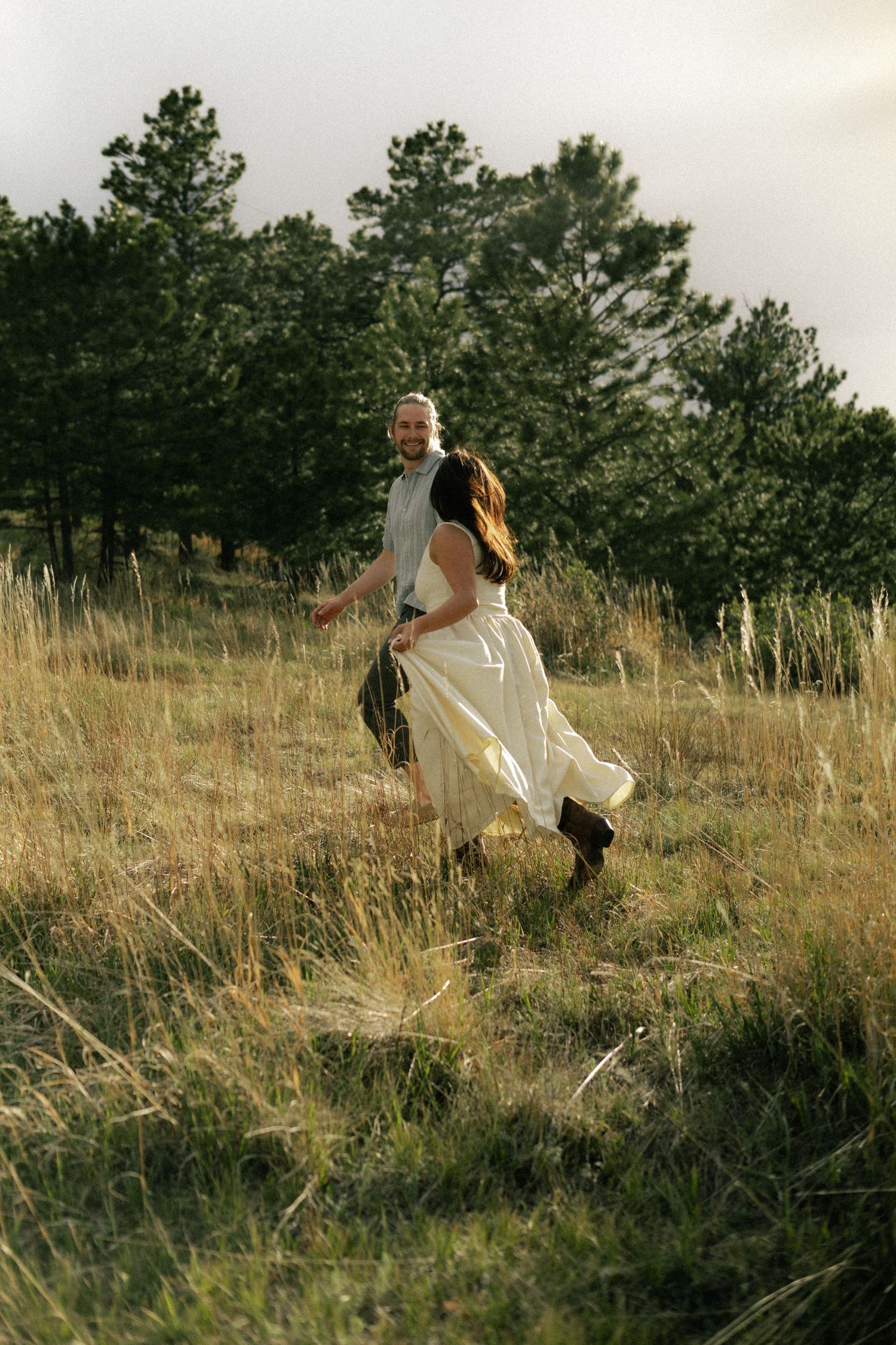 Couple running hand in hand through golden meadow with pine trees, woman in flowing white wedding dress.