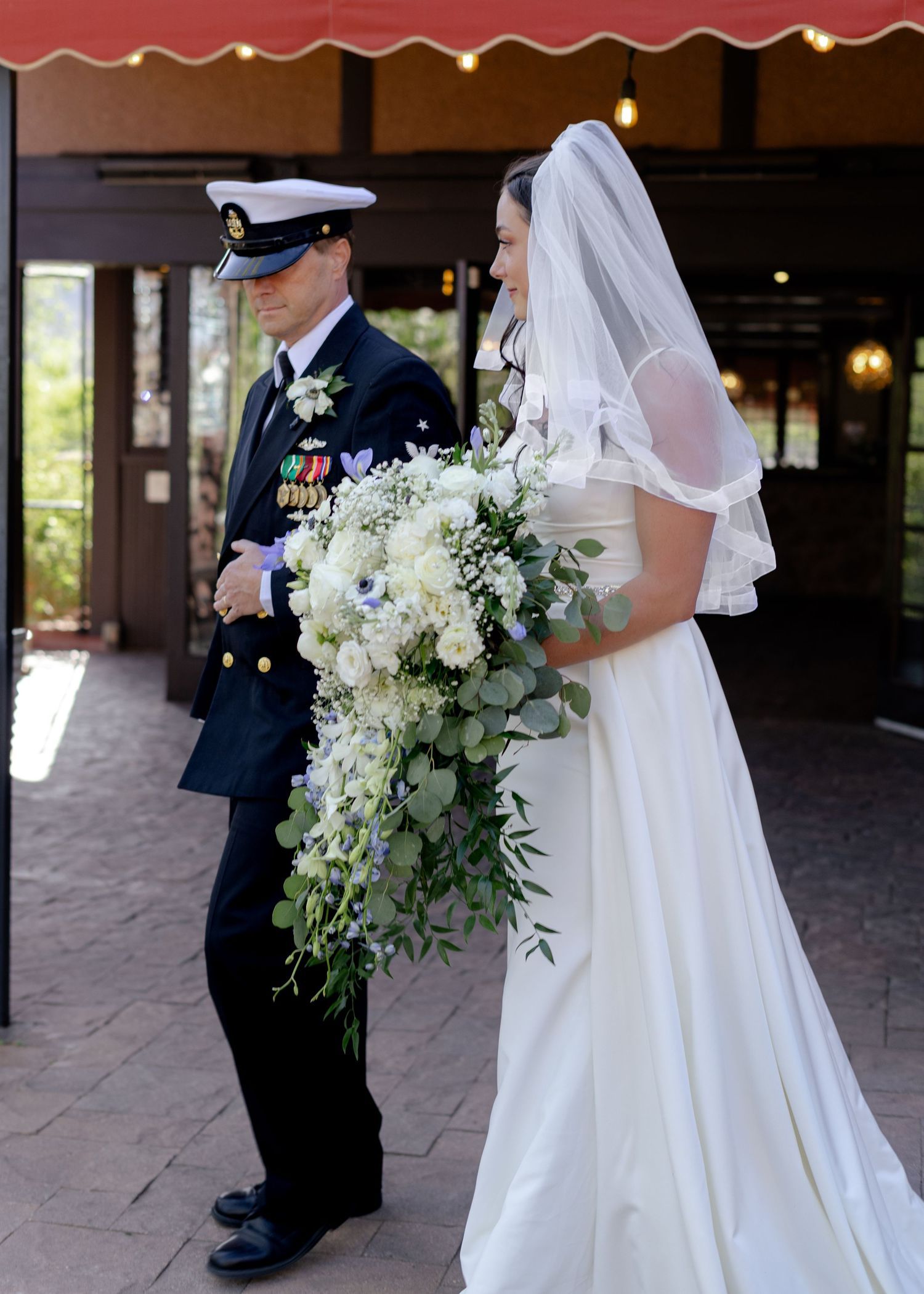 Military groom in dress uniform escorts bride in white gown and veil holding cascading white floral bouquet.
