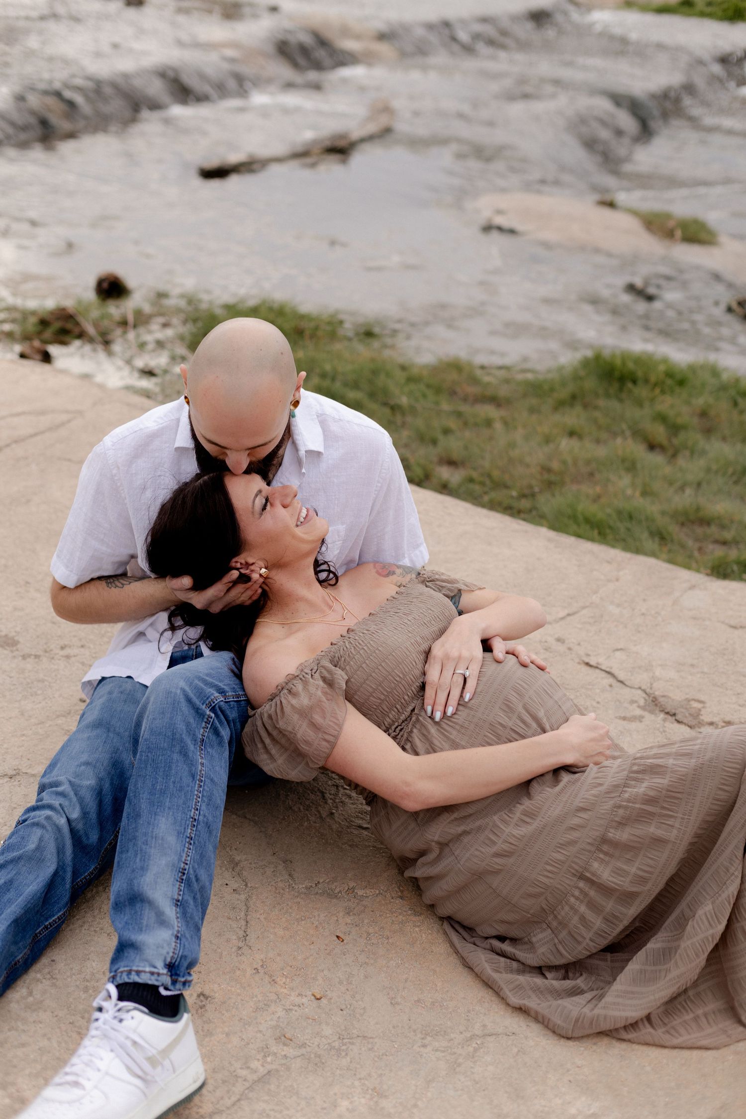 A pregnant woman in a brown dress reclines against a bald man in white shirt and jeans outdoors near rocky terrain.