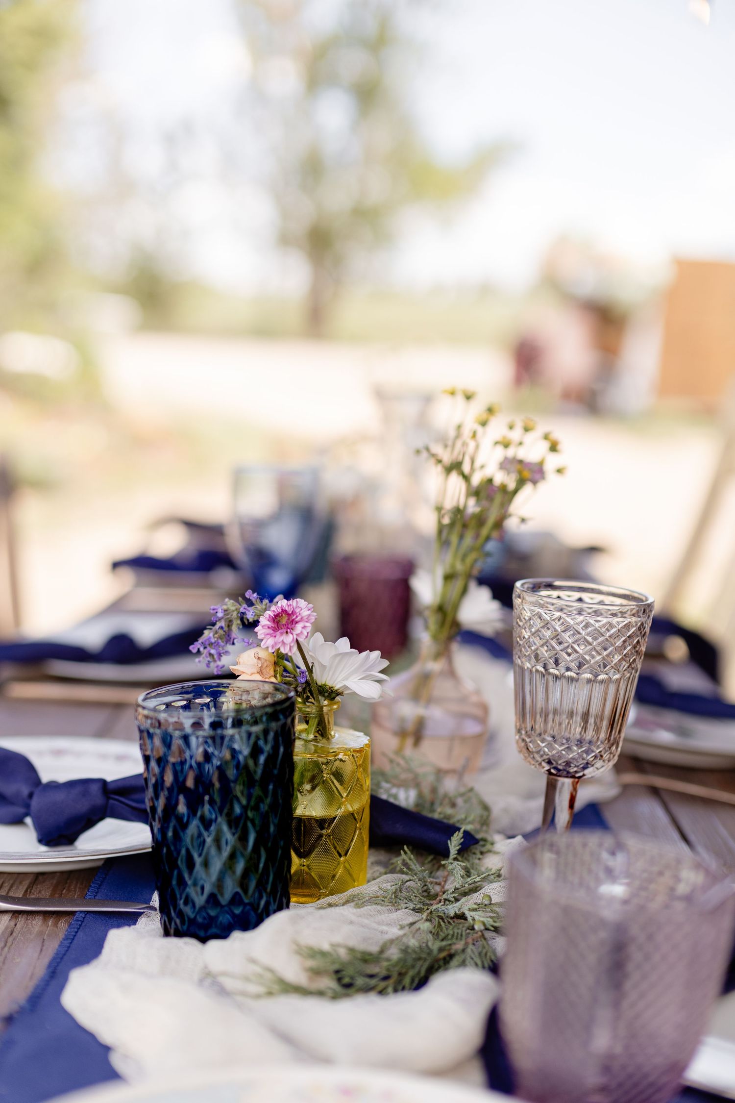 Elegant outdoor wedding table setting with colorful glassware, wildflowers, and blue linen napkins on a rustic wooden table.