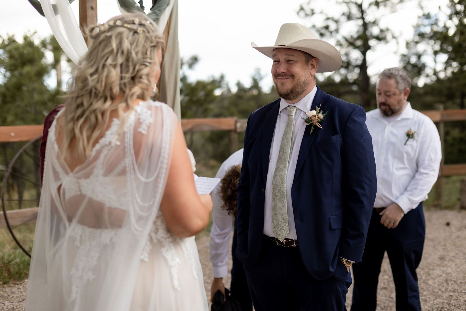 Groom in navy suit and cowboy hat smiling at bride during outdoor wedding ceremony.