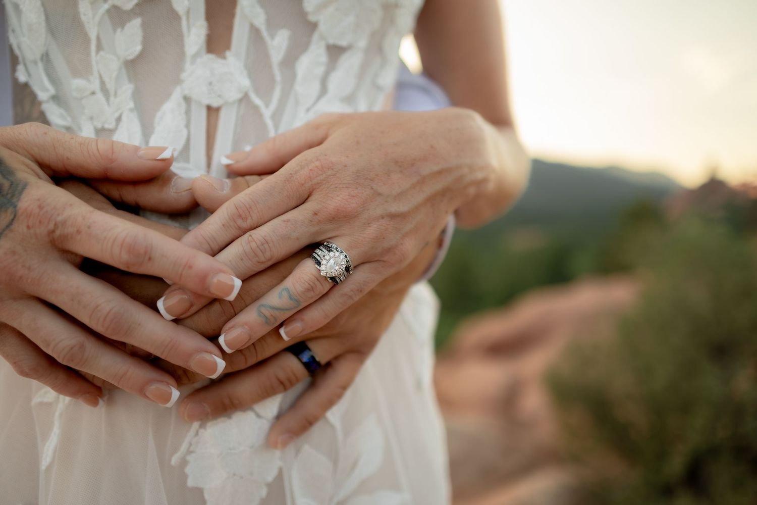 Close-up of bride and groom hands with diamond engagement ring and wedding band on white lace dress.