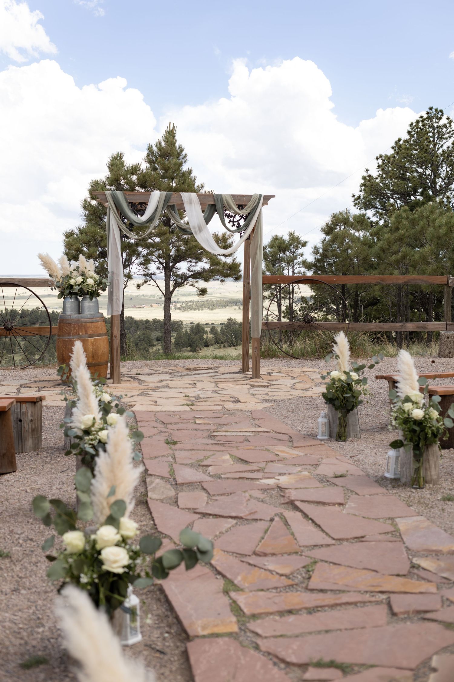 Rustic outdoor wedding ceremony setup with stone aisle, floral arrangements, and wooden arch draped with white fabric.