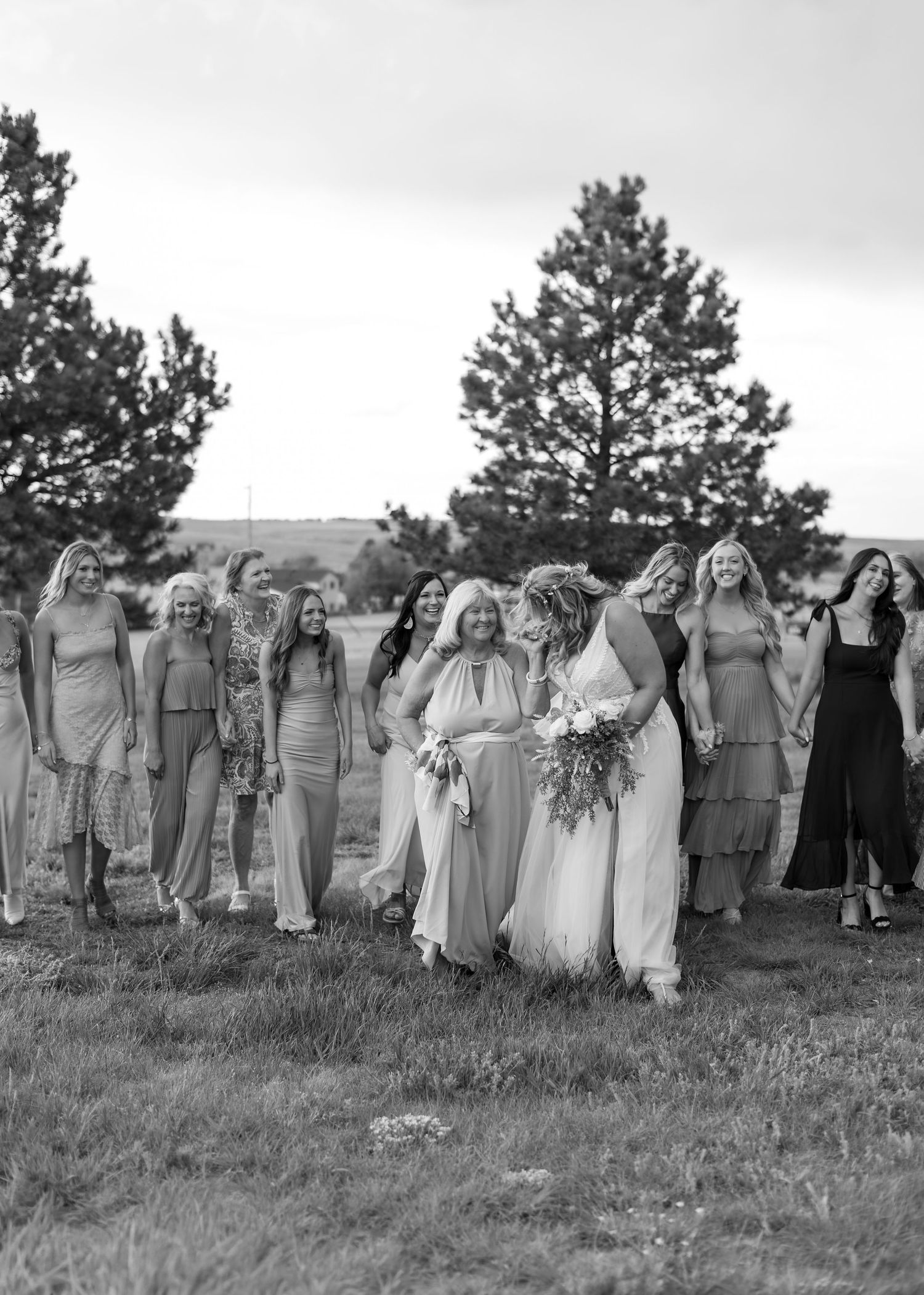 Black and white photo of a bride walking with her bridesmaids in a field with trees in the background.