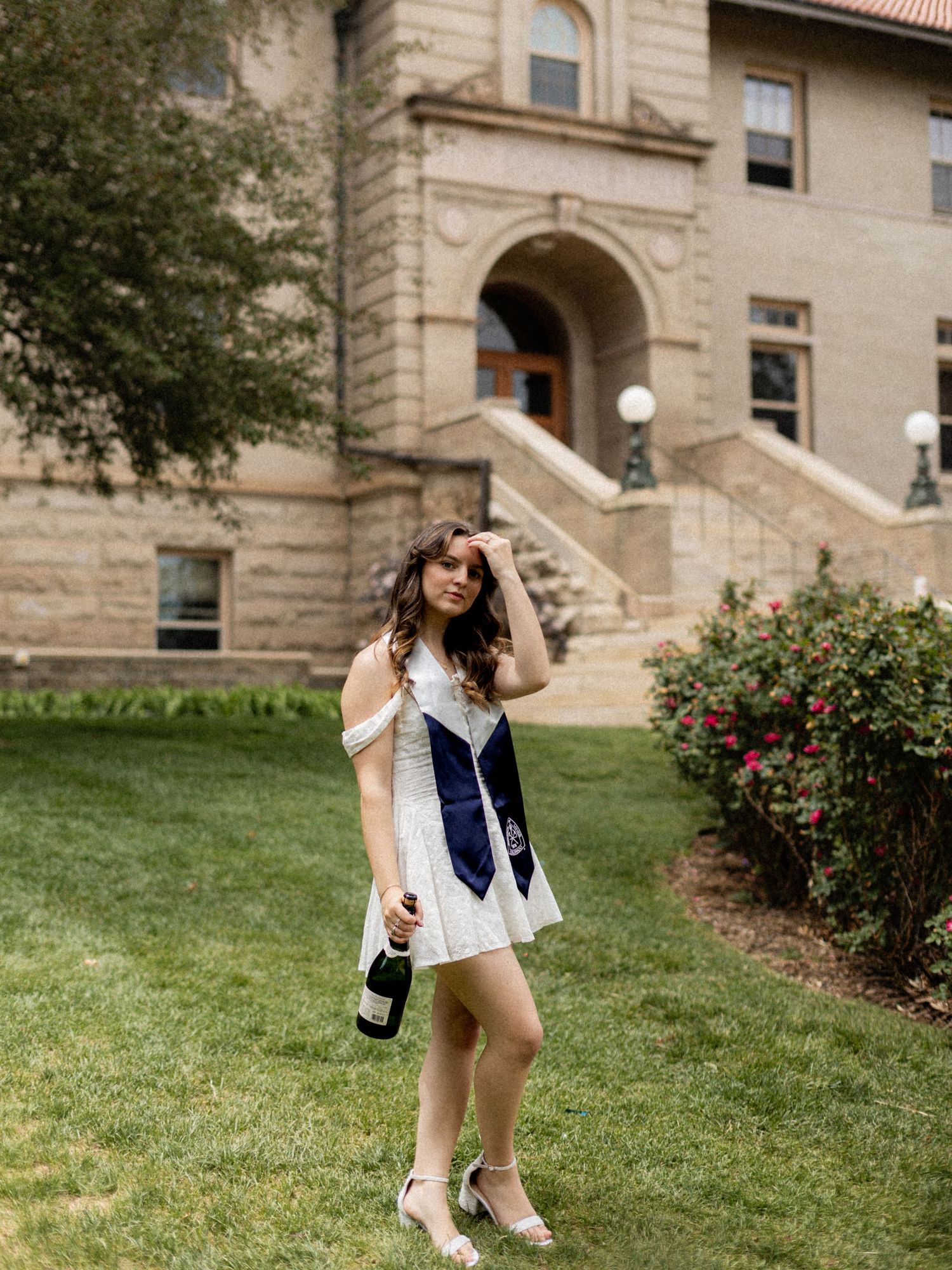 Young woman in white dress with graduation sash holds champagne bottle outside historic stone university building.