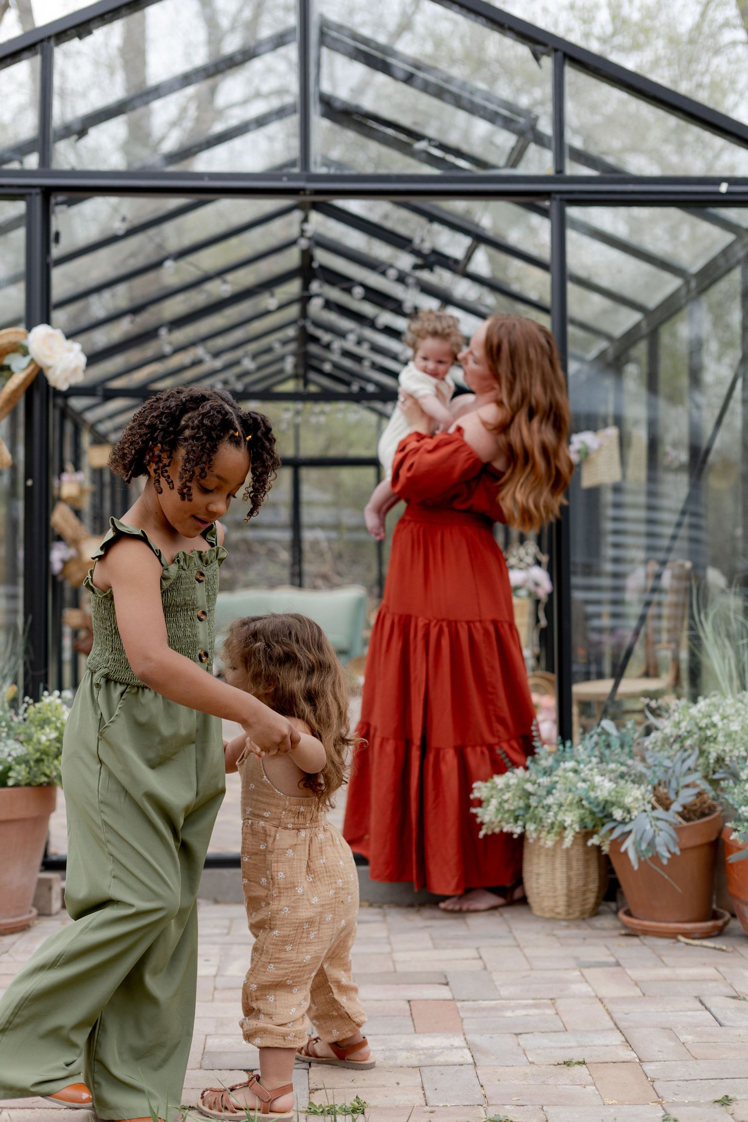 Woman in red dress holds baby near greenhouse while two young girls in green and tan jumpsuits play on brick patio.