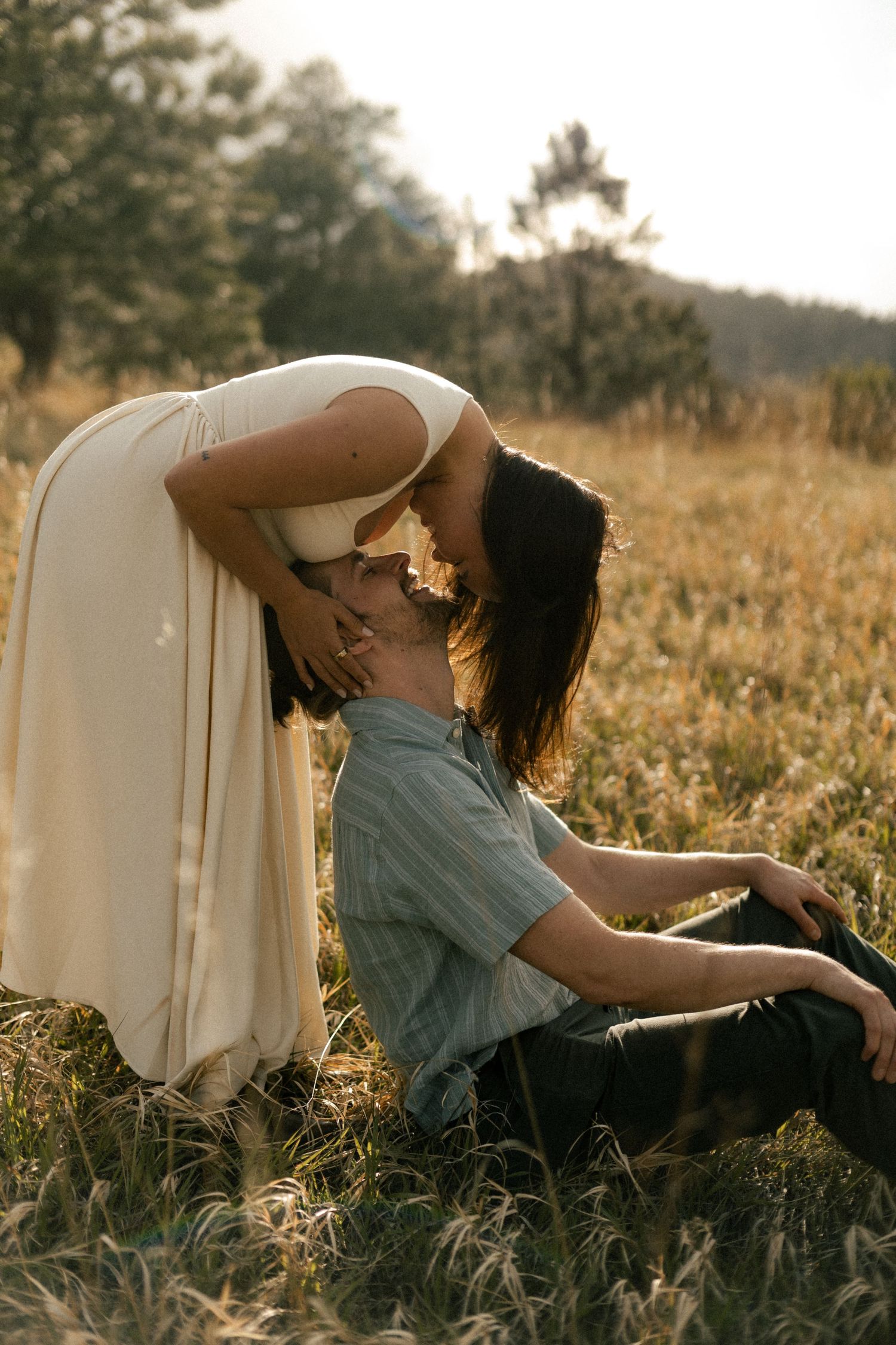 A couple shares a romantic kiss in a sunlit meadow, woman in white dress leaning over man seated in golden grass.