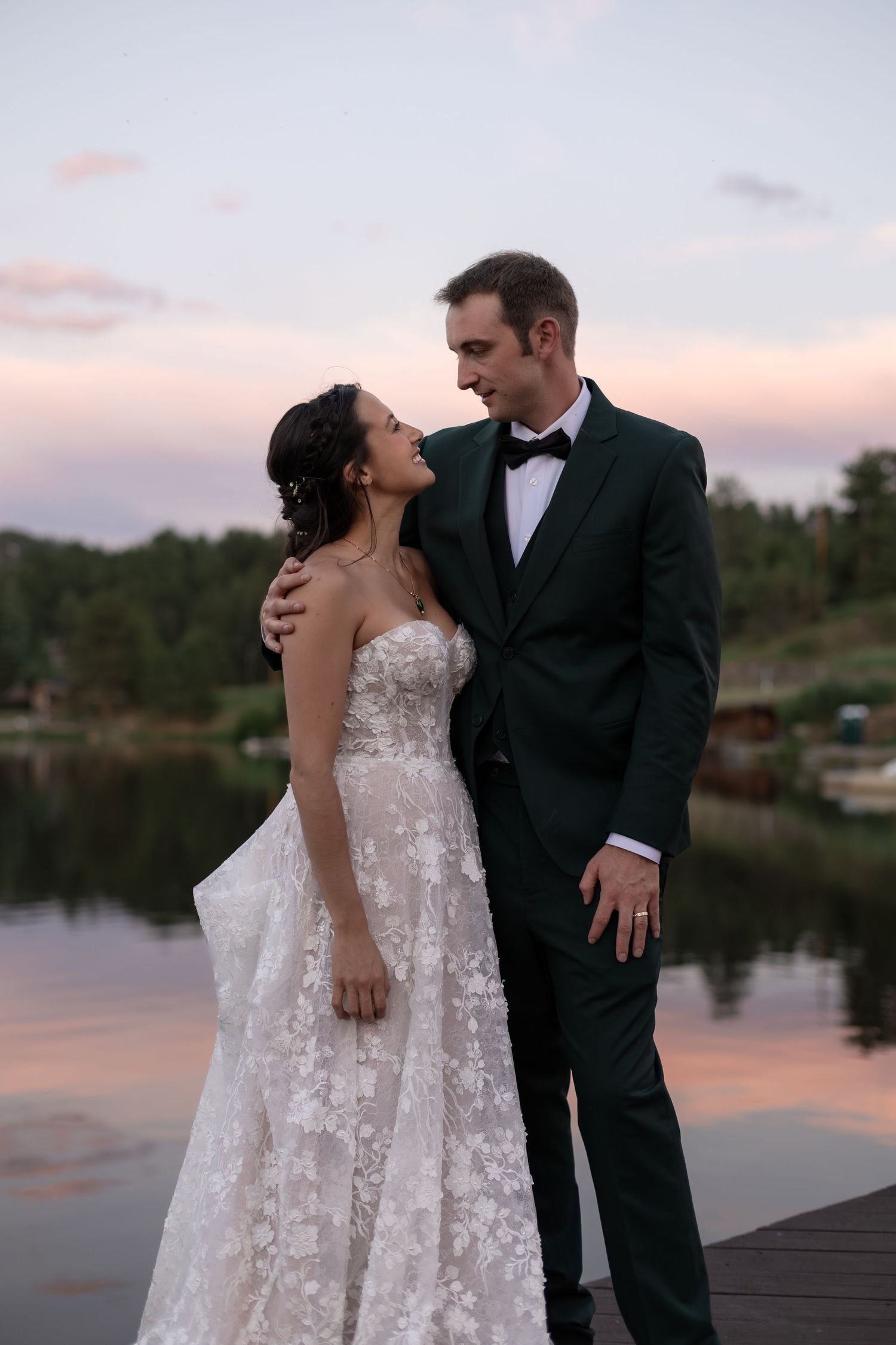 Bride and groom share a romantic moment on a dock at sunset, with a lake and pink sky in the background.