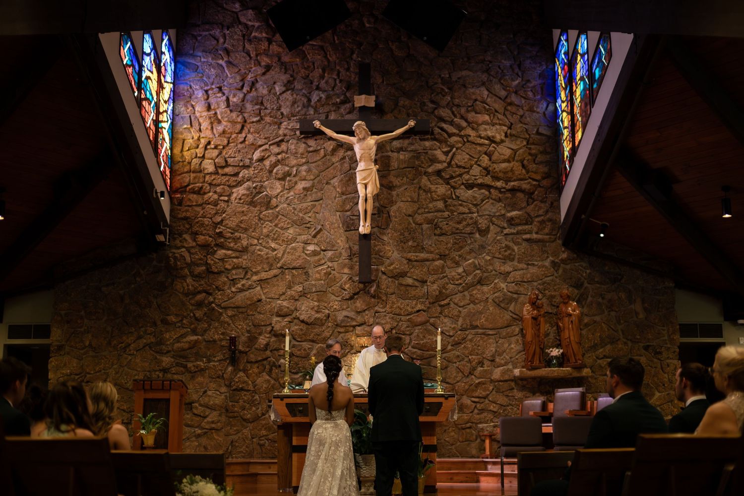 Bride and groom stand before a stone altar with crucifix during a Catholic wedding ceremony in a chapel.