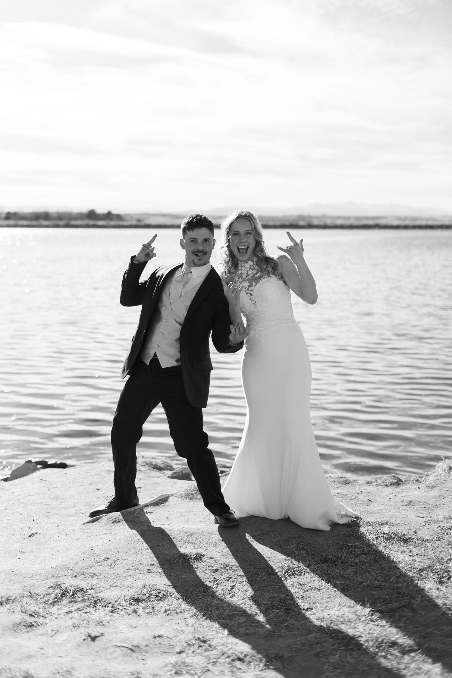 Black and white photo of bride and groom making rock on gestures by a lake on their wedding day.