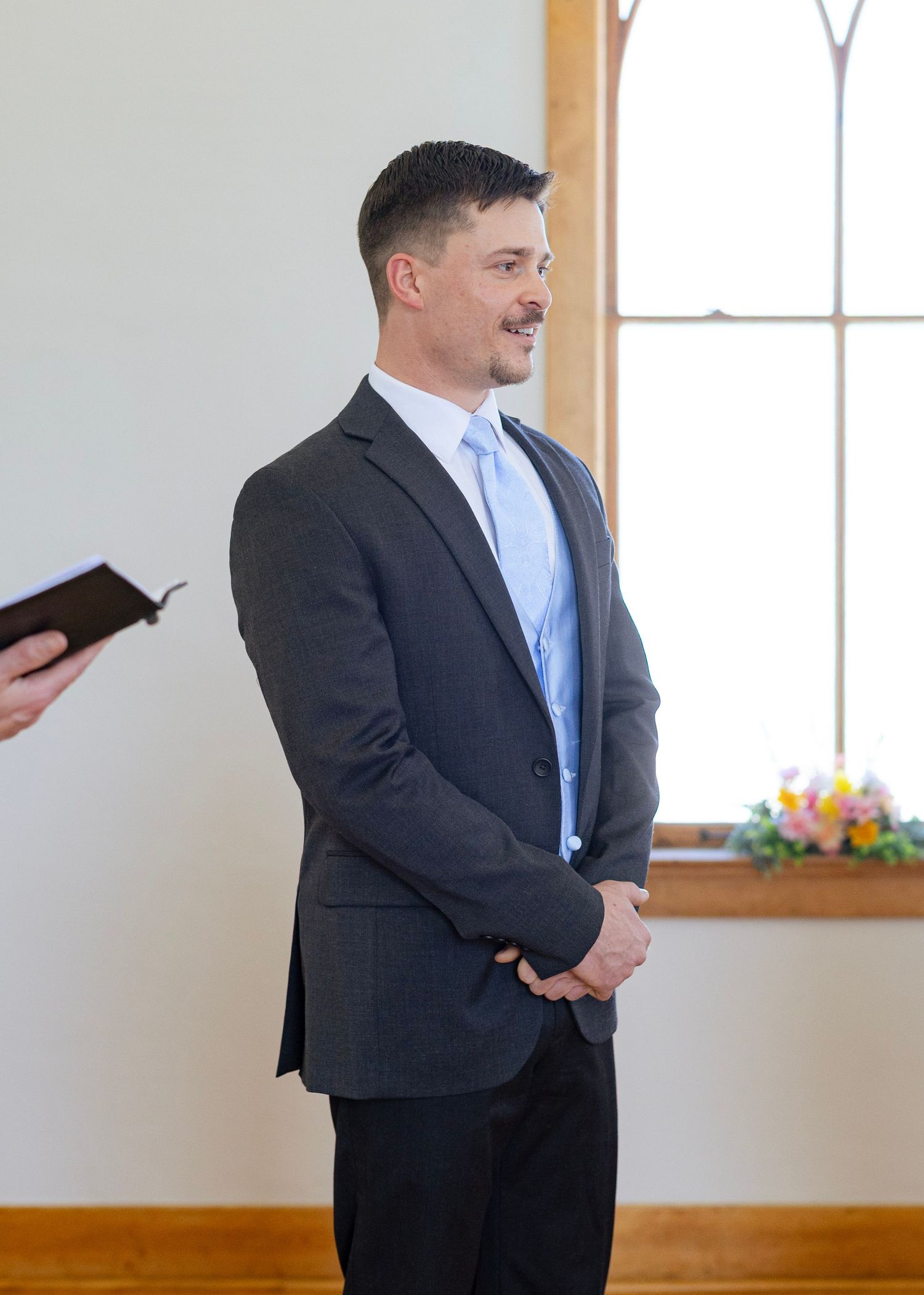 A man in a dark gray suit and light blue tie smiles while standing at a wedding ceremony altar.