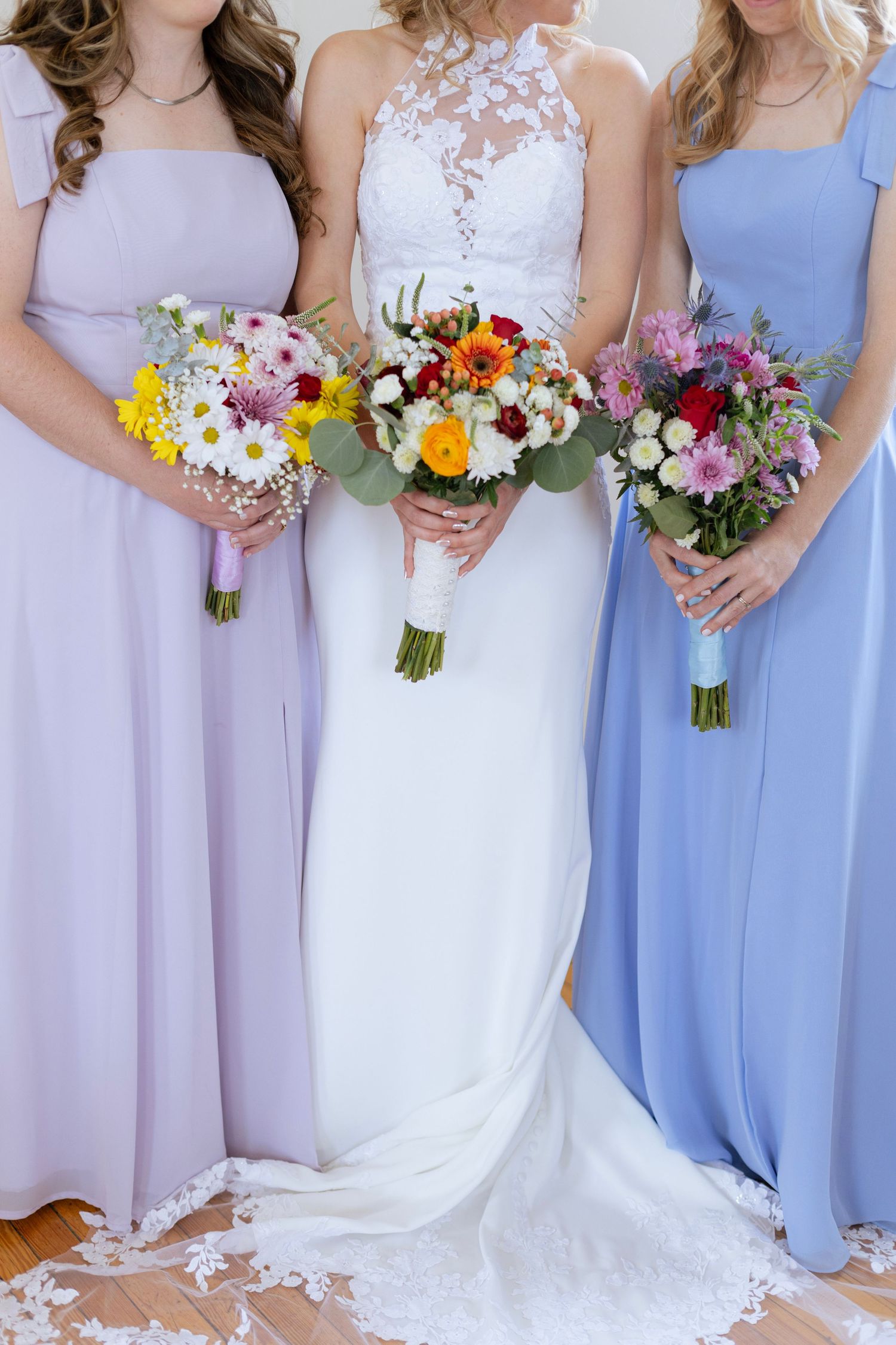 Bride in lace halter gown flanked by bridesmaids in lavender and blue dresses holding colorful flower bouquets.