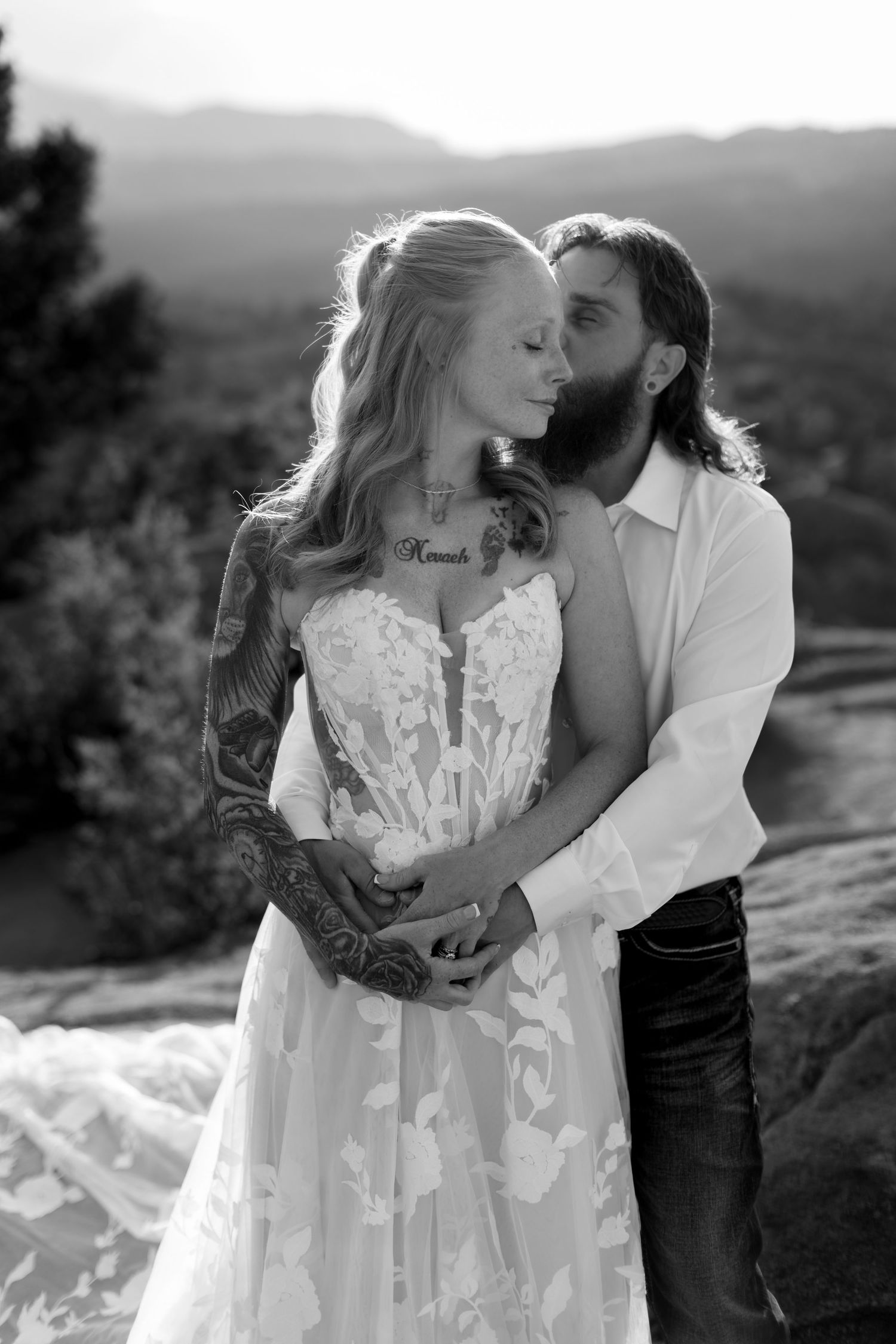 Black and white photo of a tattooed bride in lace gown embraced by bearded groom against mountain backdrop.