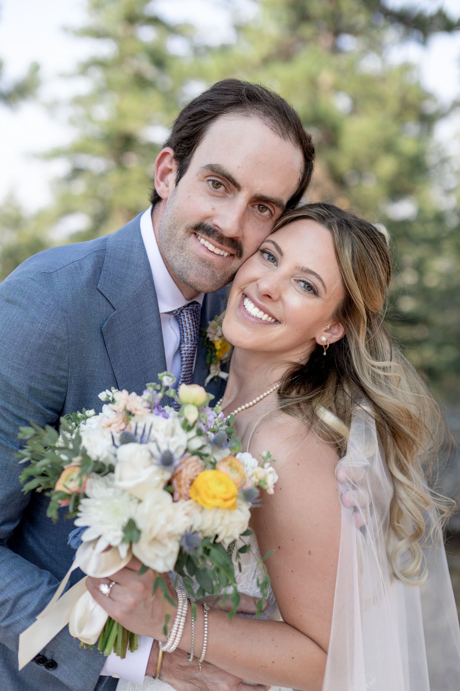 Happy bride and groom smiling together on their wedding day, bride holding a beautiful bouquet of white, yellow, and purple flowers.
