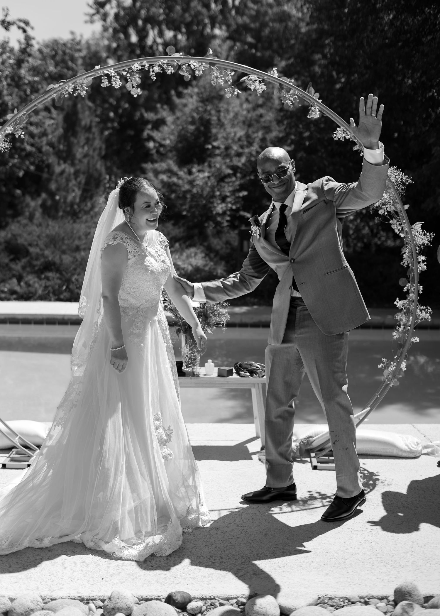 Black and white photo of a joyful bride and groom celebrating under a floral arch near a pool.