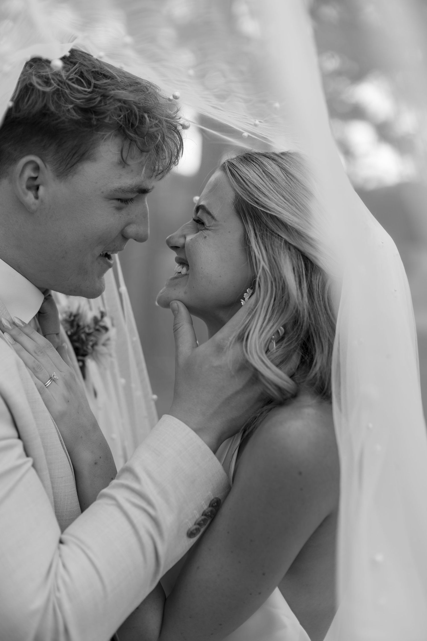Black and white wedding portrait of smiling bride and groom sharing a tender moment under veil.
