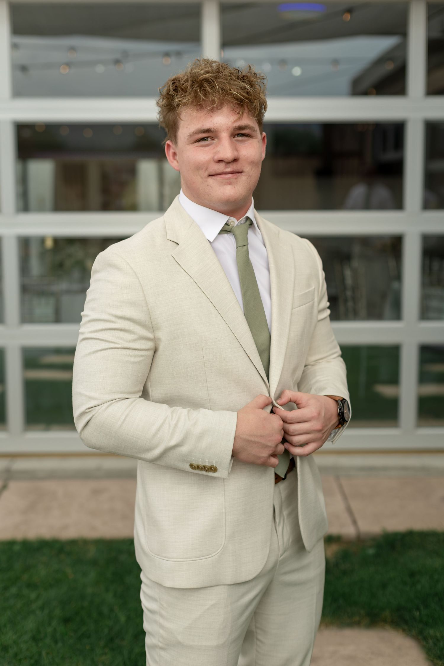 Young groom in cream suit and sage green tie buttons jacket while posing outdoors before wedding.
