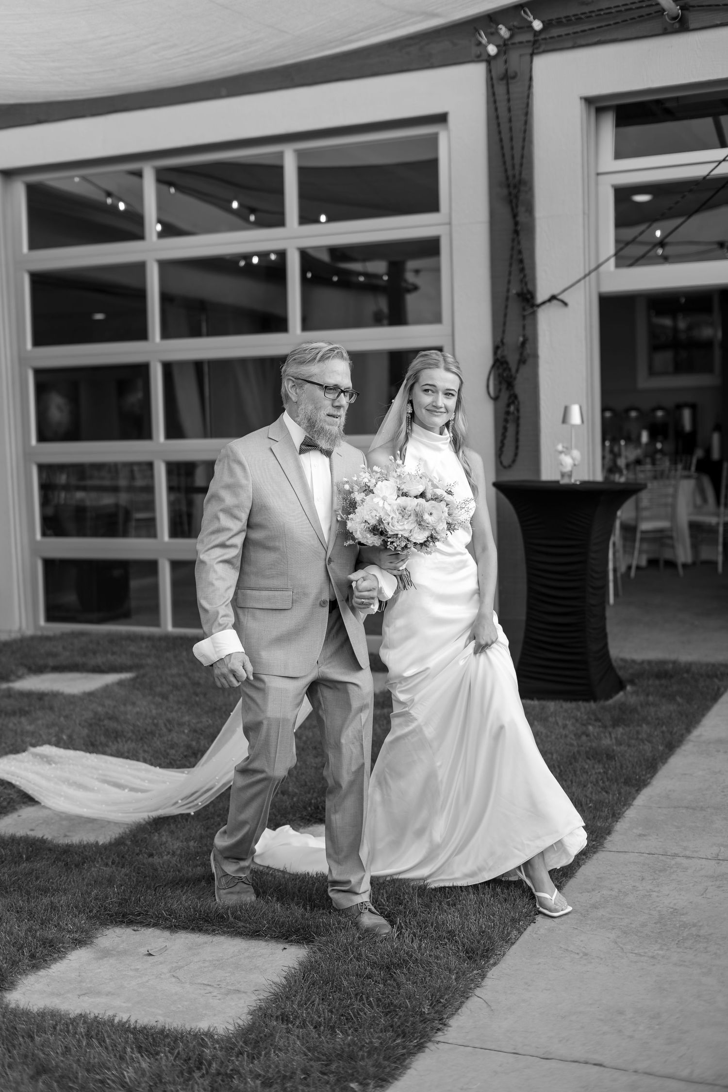 Black and white photo of a bride in a white gown and veil walking with an older man in a suit at an outdoor wedding.