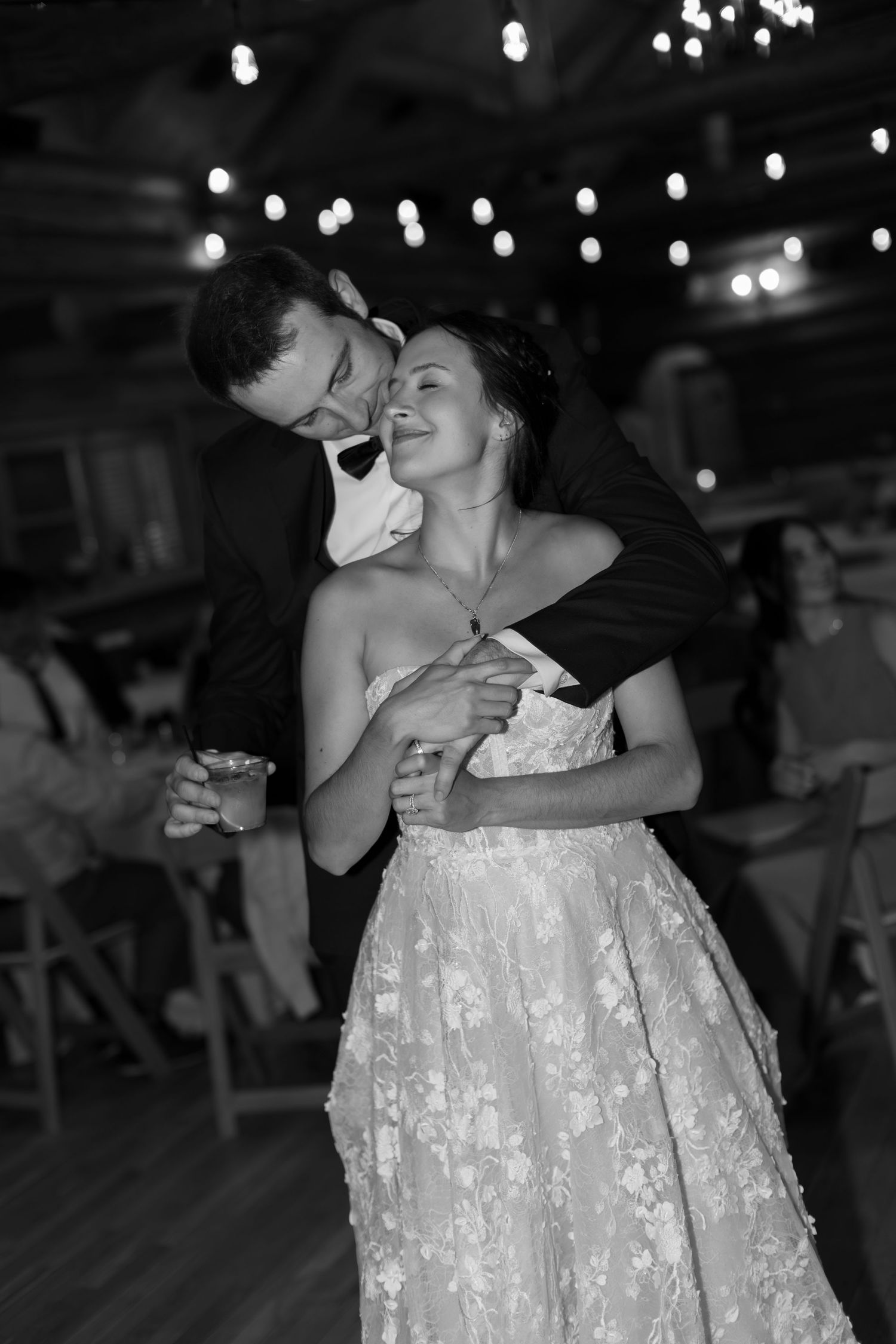 Black and white photo of a bride and groom sharing their first dance, embracing romantically under string lights.