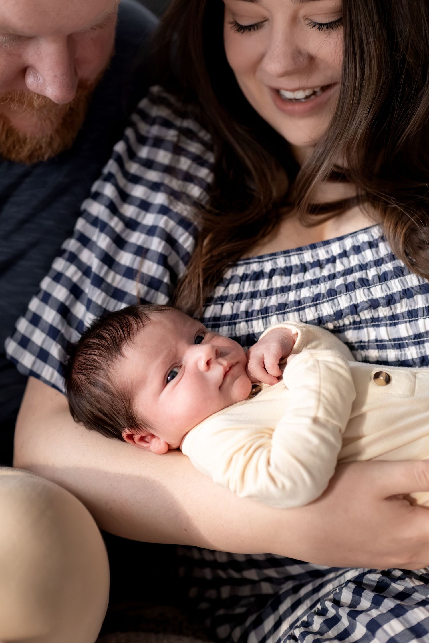 New parents smiling and cradling their newborn baby dressed in a white onesie during an intimate family portrait.