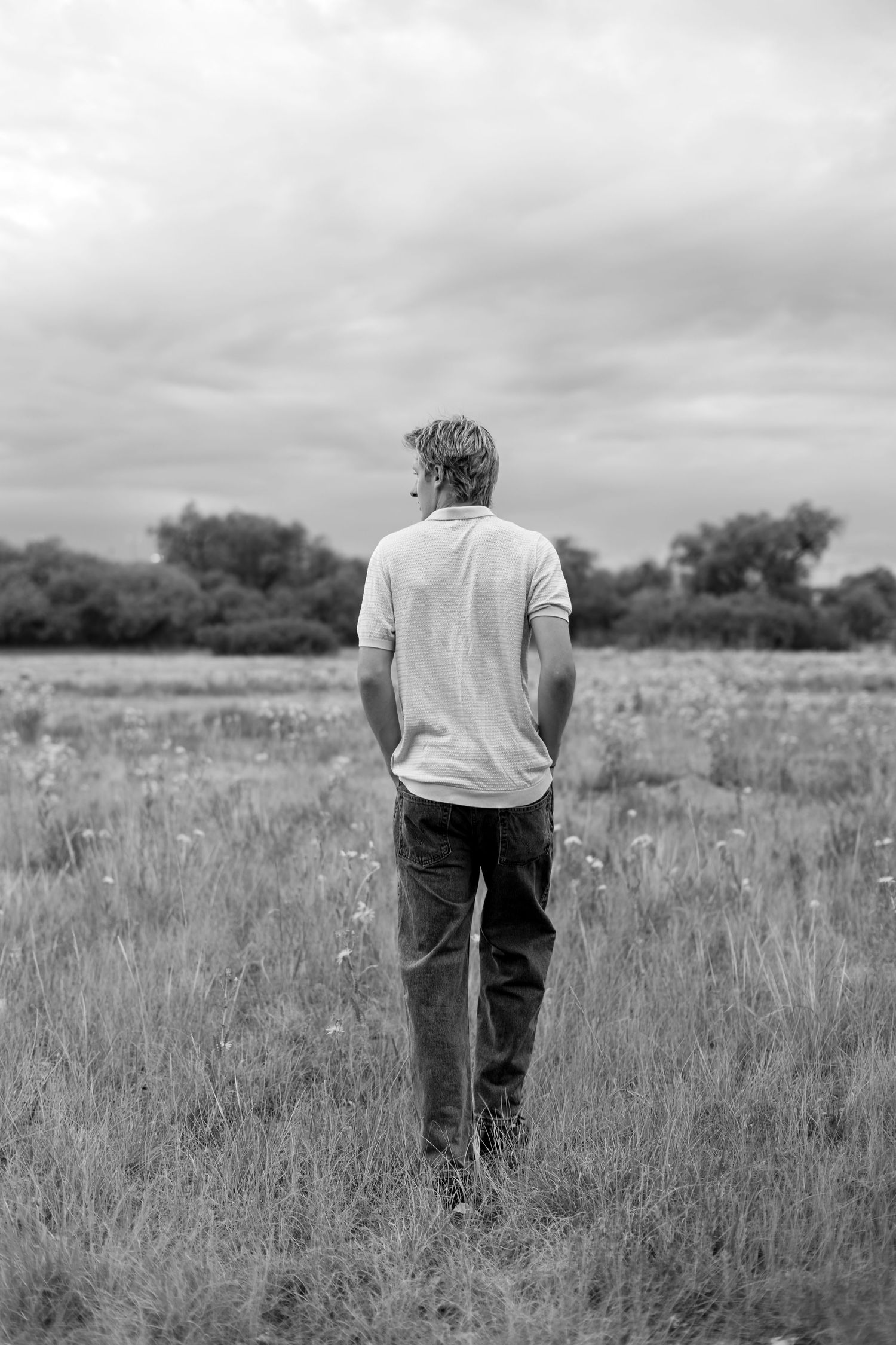 Black and white photo of a man standing alone in a field, gazing at a cloudy sky with trees in the background.