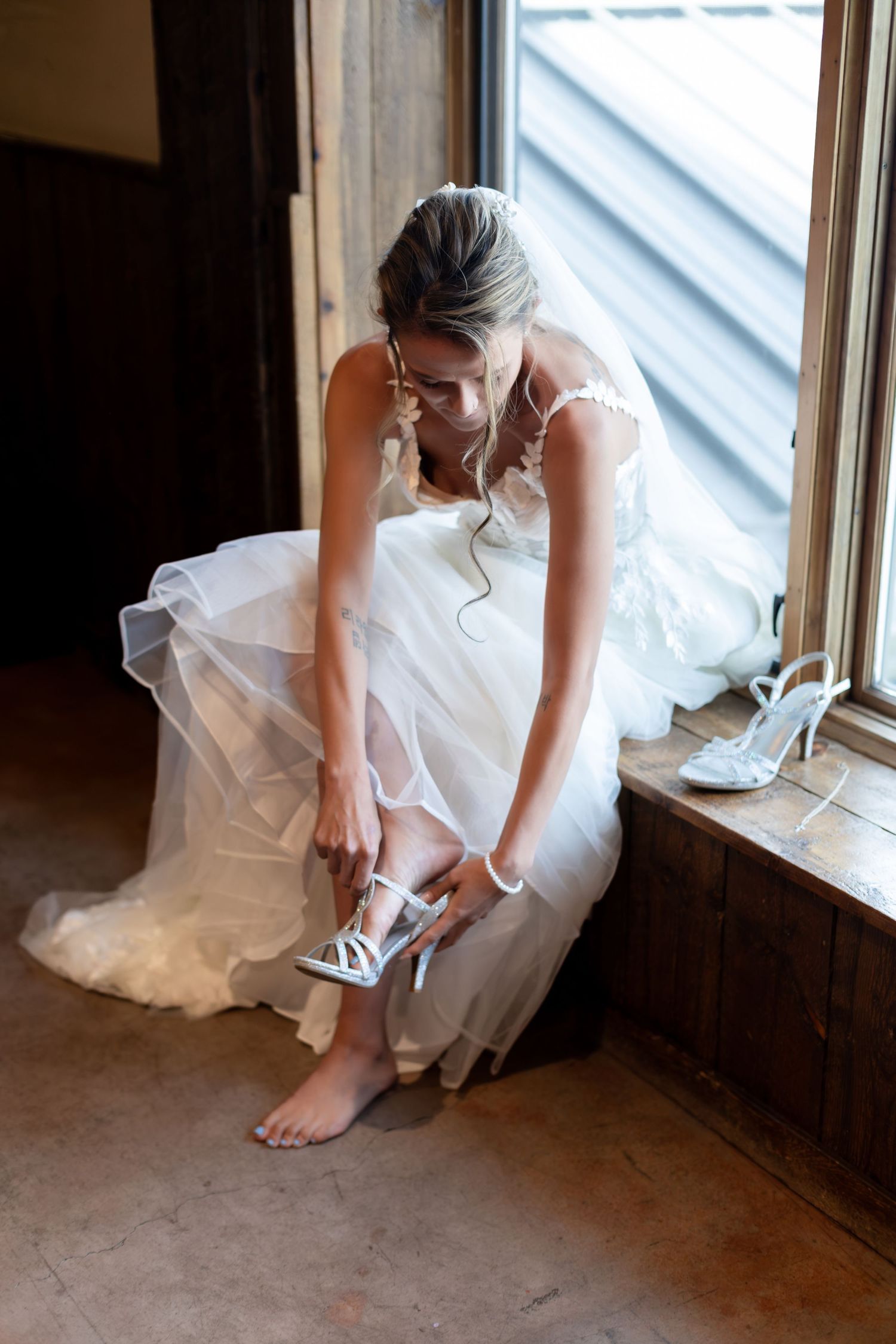 Bride in white wedding gown putting on silver heeled shoes while seated by rustic barn window.