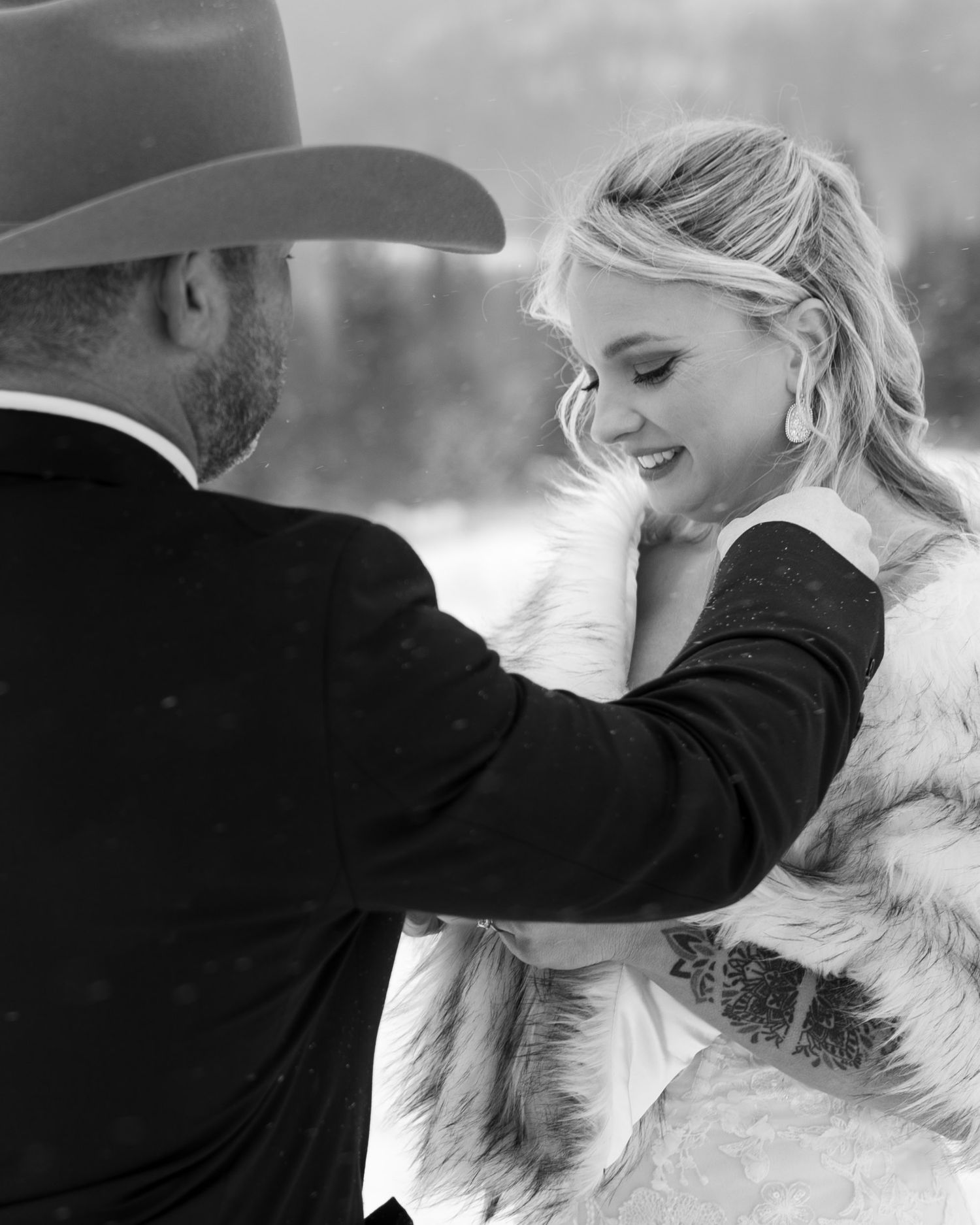 Black and white winter wedding photo of bride in fur wrap smiling as groom in cowboy hat tenderly touches her face.