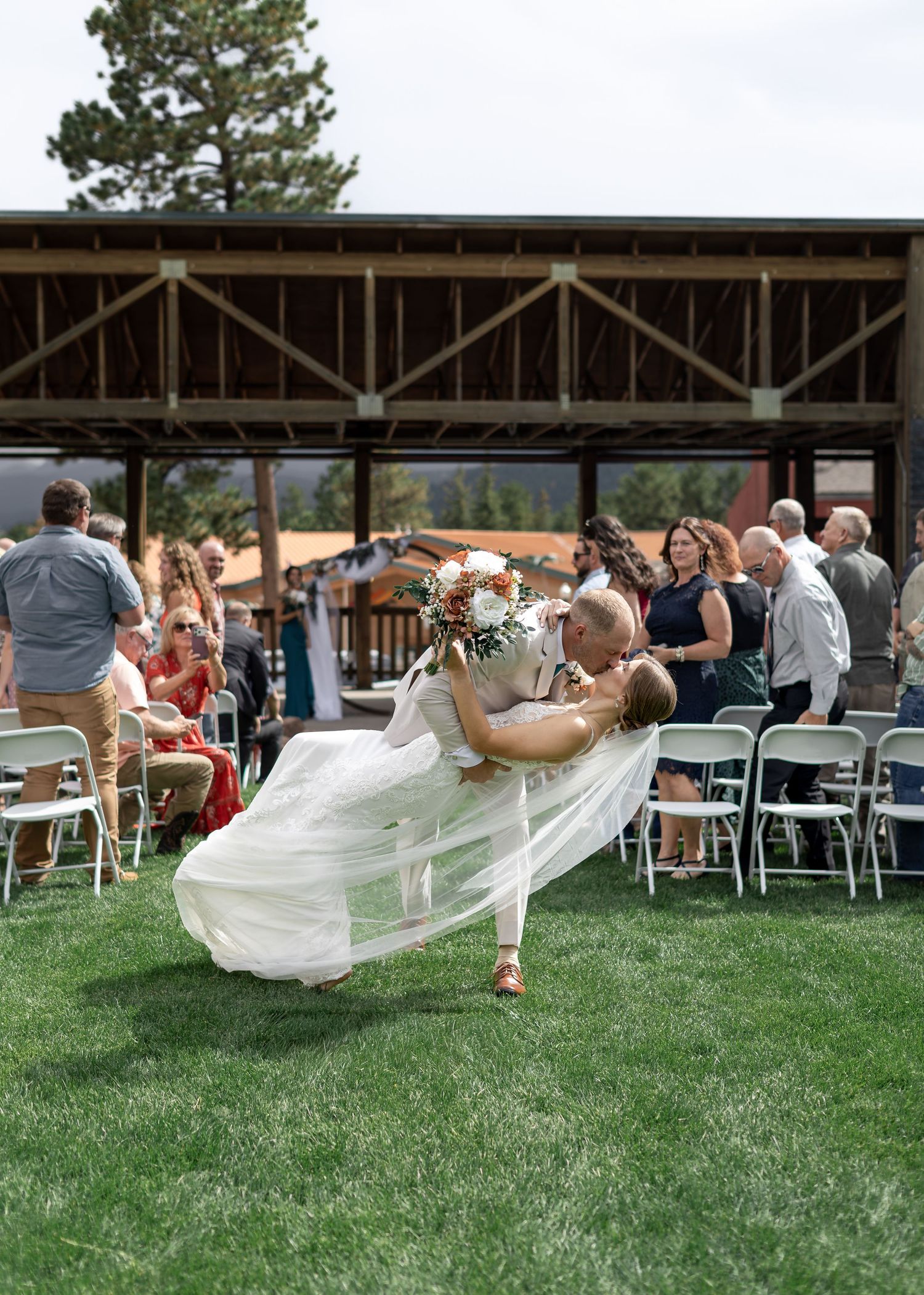Groom dips bride in white gown during outdoor wedding ceremony on green lawn with seated guests.