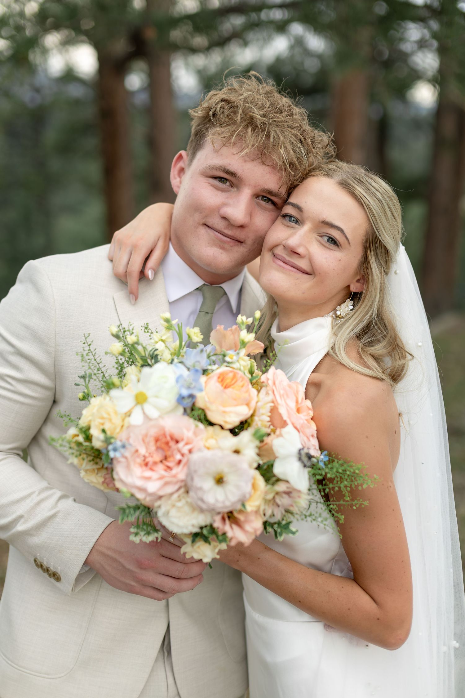 Smiling bride and groom pose outdoors holding a pastel bouquet of roses, the bride in white gown and veil.