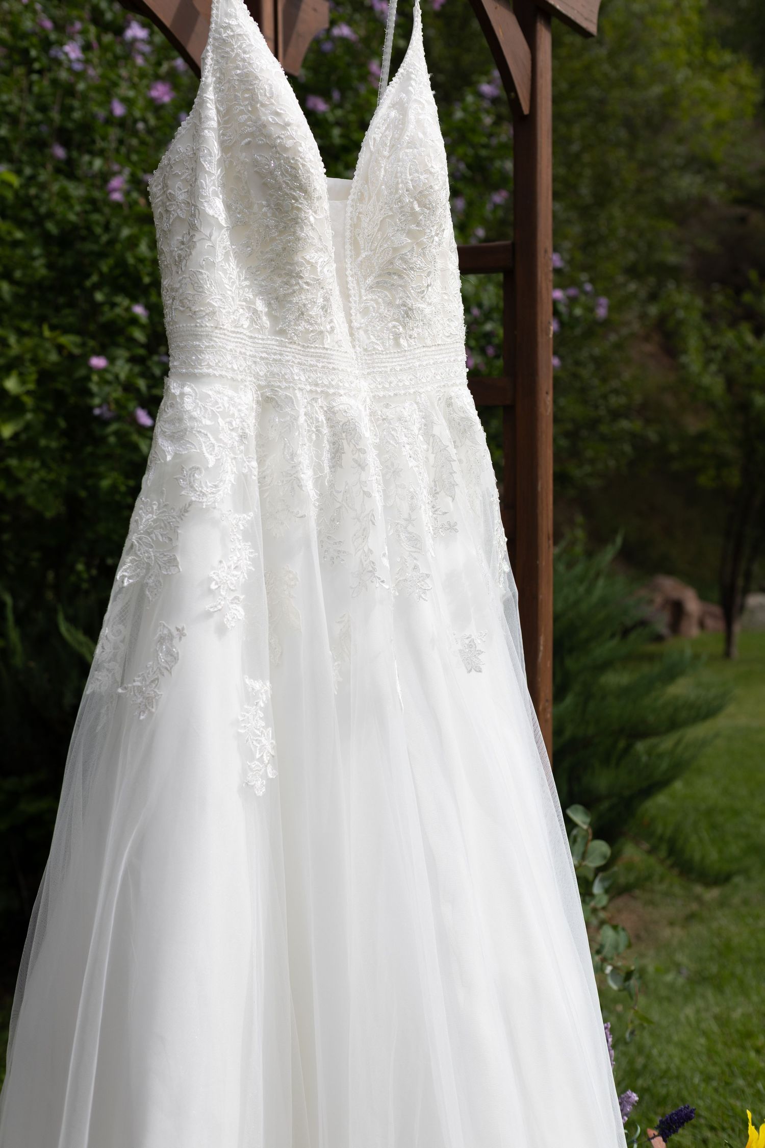 White lace A-line wedding dress with spaghetti straps hanging outdoors on a wooden arbor surrounded by greenery.