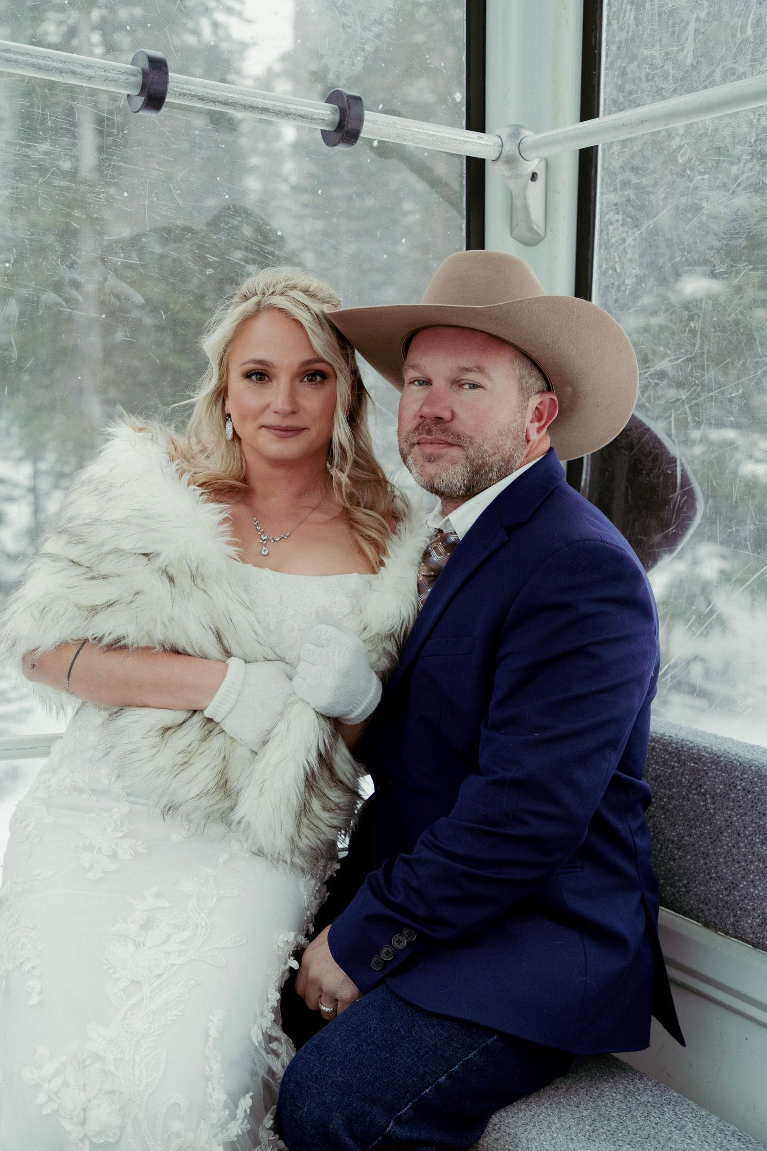 A couple poses together in a snowy winter setting wearing formal attire with a white fur wrap and navy suit.